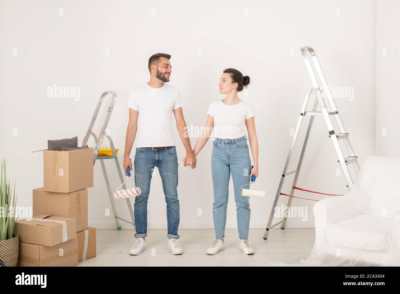 Sorridi una giovane coppia in jeans che tiene le mani e si guarda l'un l'altro mentre si sostiene durante il rimodellamento della stanza Foto Stock