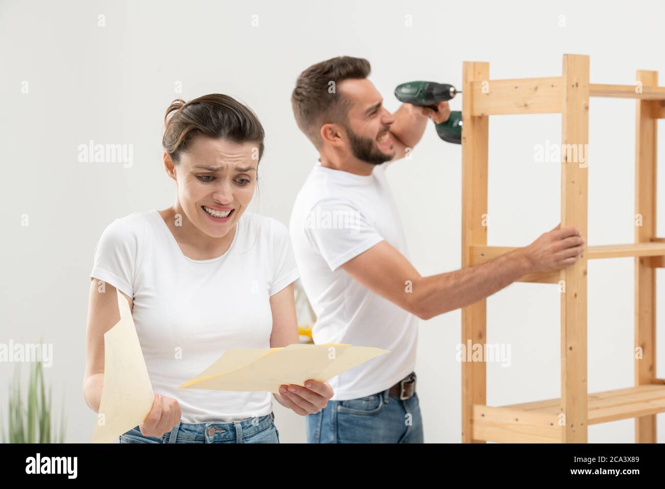 La giovane donna accigliata ha perplessato guardando il piano di assemblaggio con confusione mentre il suo marito infastidito assemblando i mobili Foto Stock