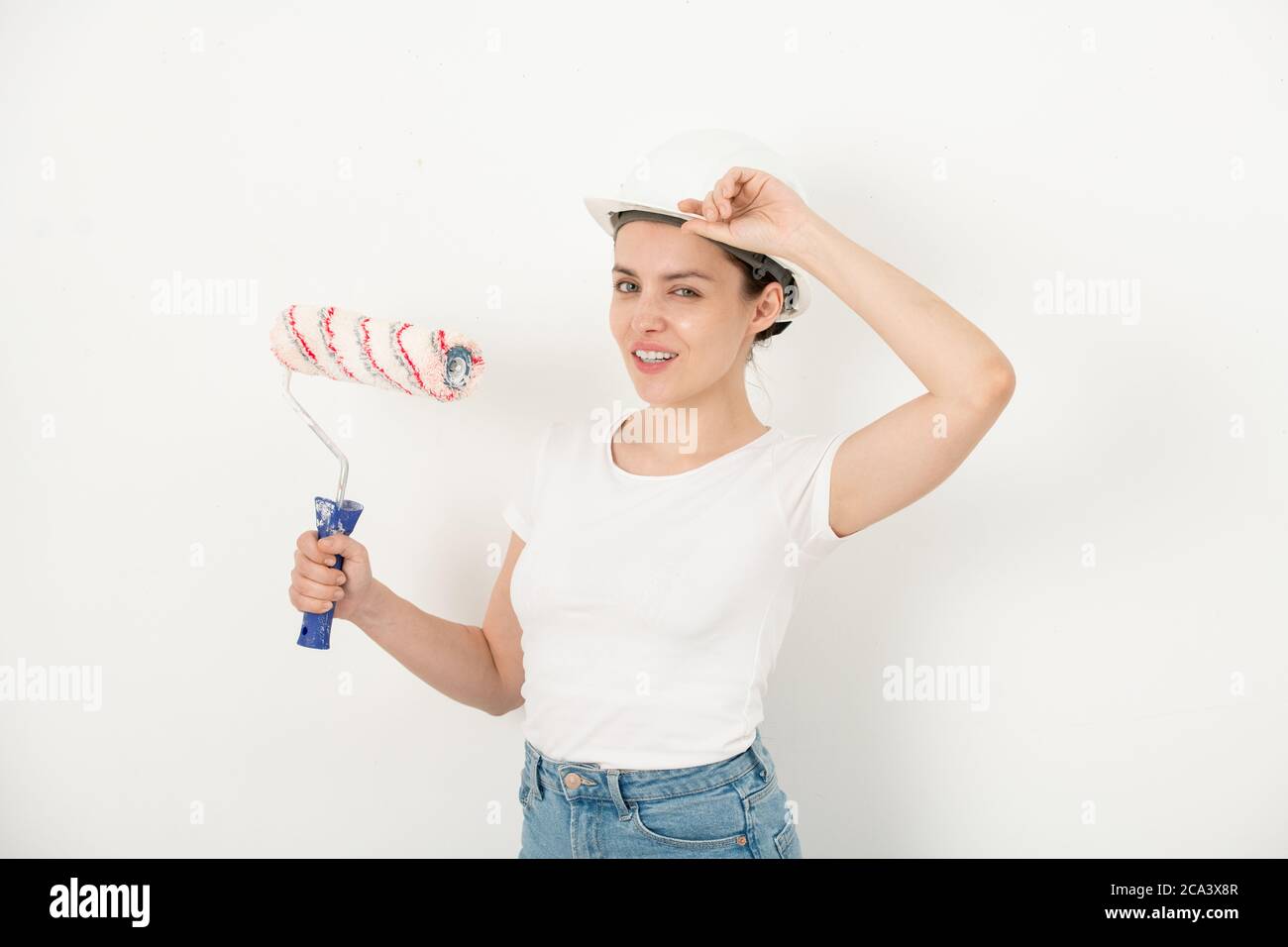 Ritratto di giovane donna sicura positiva che regola il cappello e tiene il rotolo di vernice contro la parete bianca Foto Stock