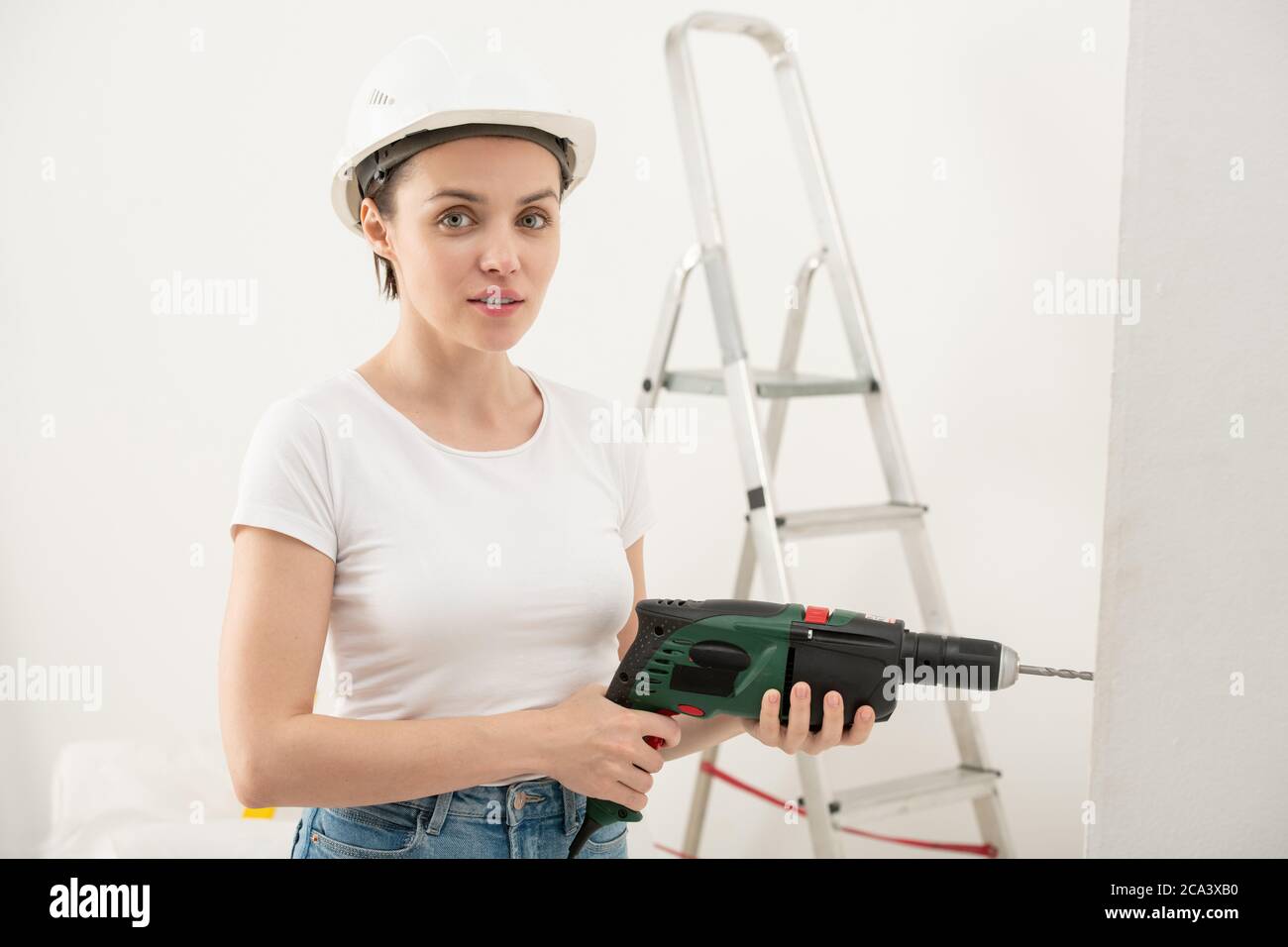 Ritratto di soddisfare giovane donna in hardhat usando il trapano elettrico mentre facendo la ristrutturazione in nuovo appartamento Foto Stock