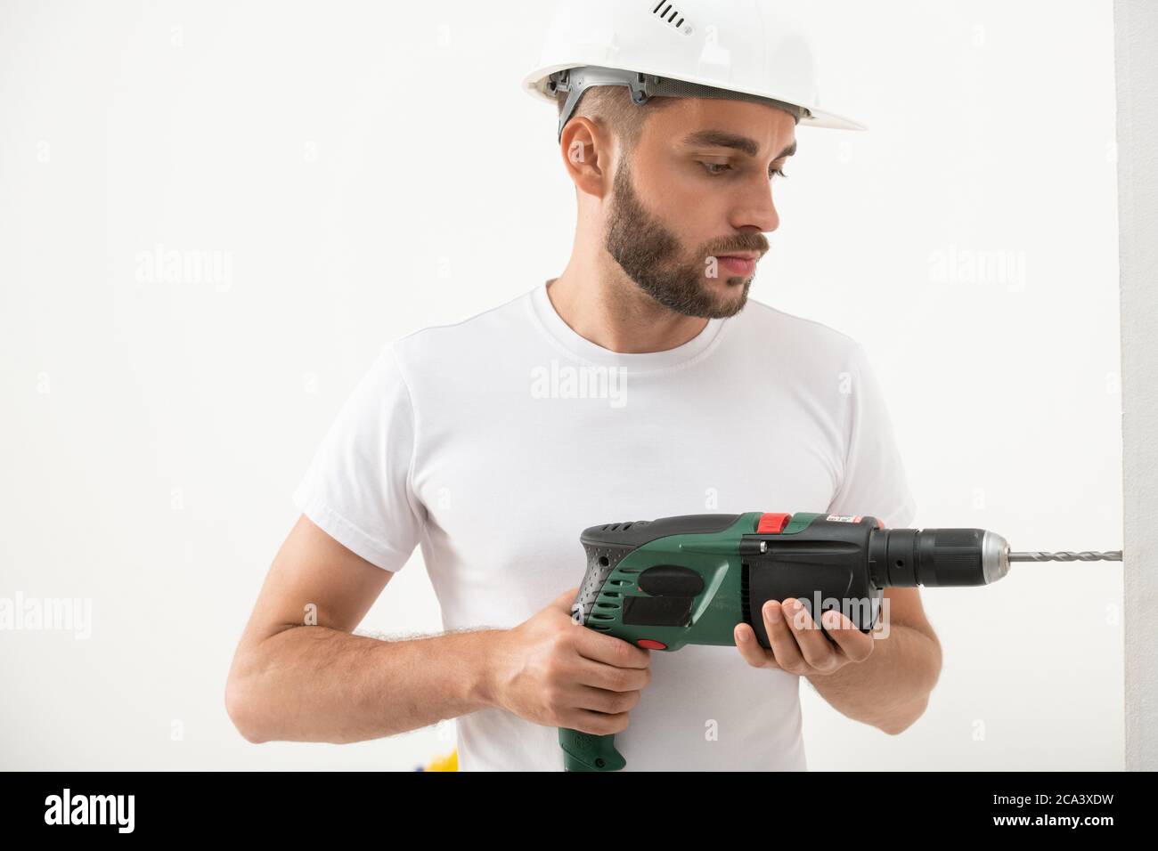 Concentrato giovane uomo bearded in costruzione hardhat che perfora il foro nella parete mentre facendo la ristrutturazione in nuovo piano Foto Stock