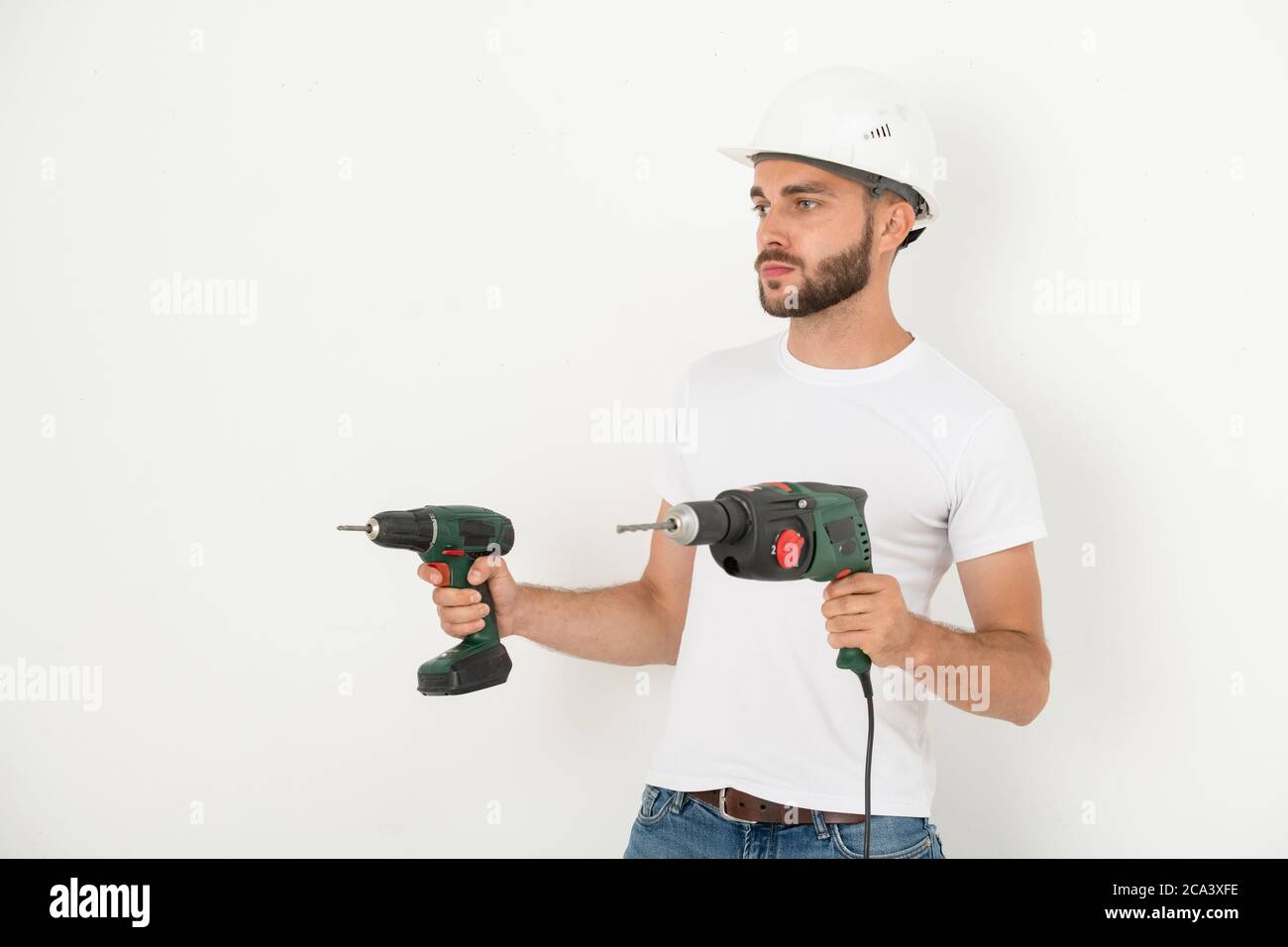 Riparatore giovane e serio in hardhat che tiene gli attrezzi manuali di potere mentre si prepara per rimodellamento in piano Foto Stock