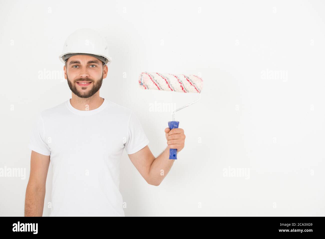 Ritratto di sorridente bel giovane decoratore maschio con stoppia indossando hardhat tenendo rullo di vernice contro parete bianca Foto Stock