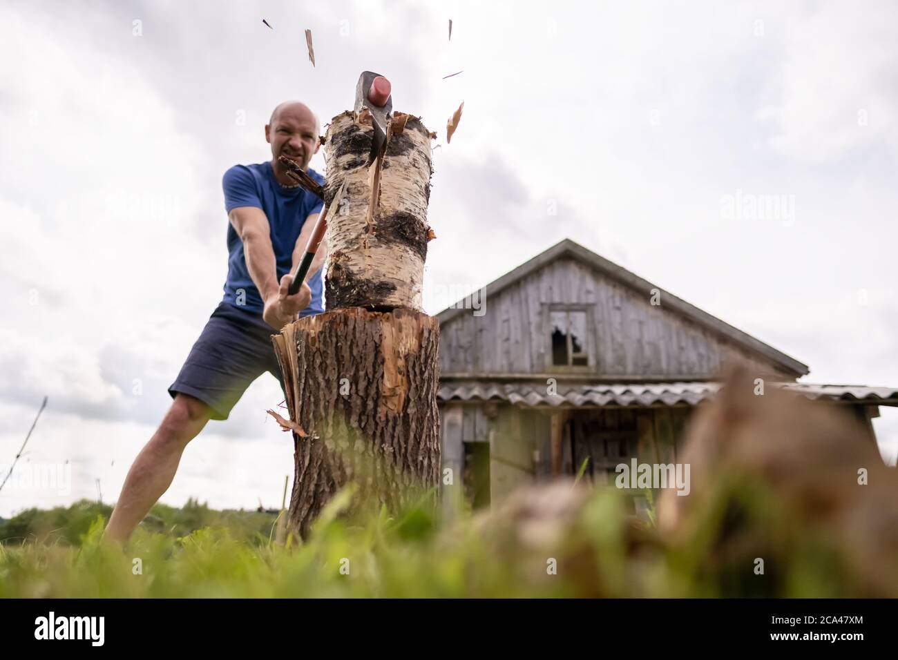 Uomo forte lumberjack tritare legno per l'inverno, con una grande ascia d'acciaio, nel villaggio, sullo sfondo di un vecchio fienile. Foto Stock