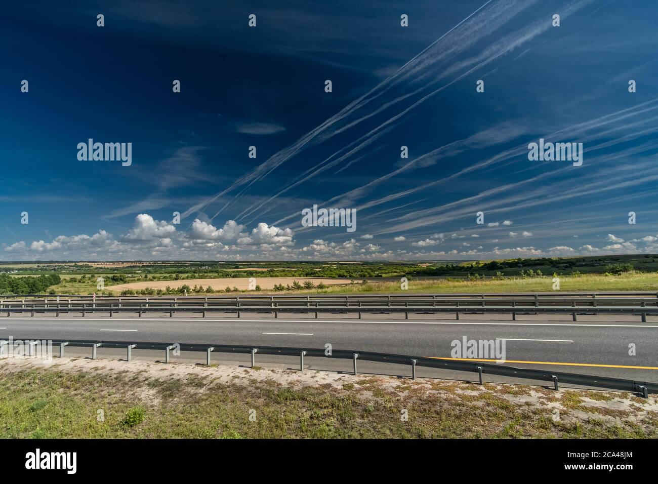 Tracce bianche di condensa di vapore acqueo sotto forma di linee da aerei volanti sopra l'autostrada, sul campo e gli alberi. Foto Stock