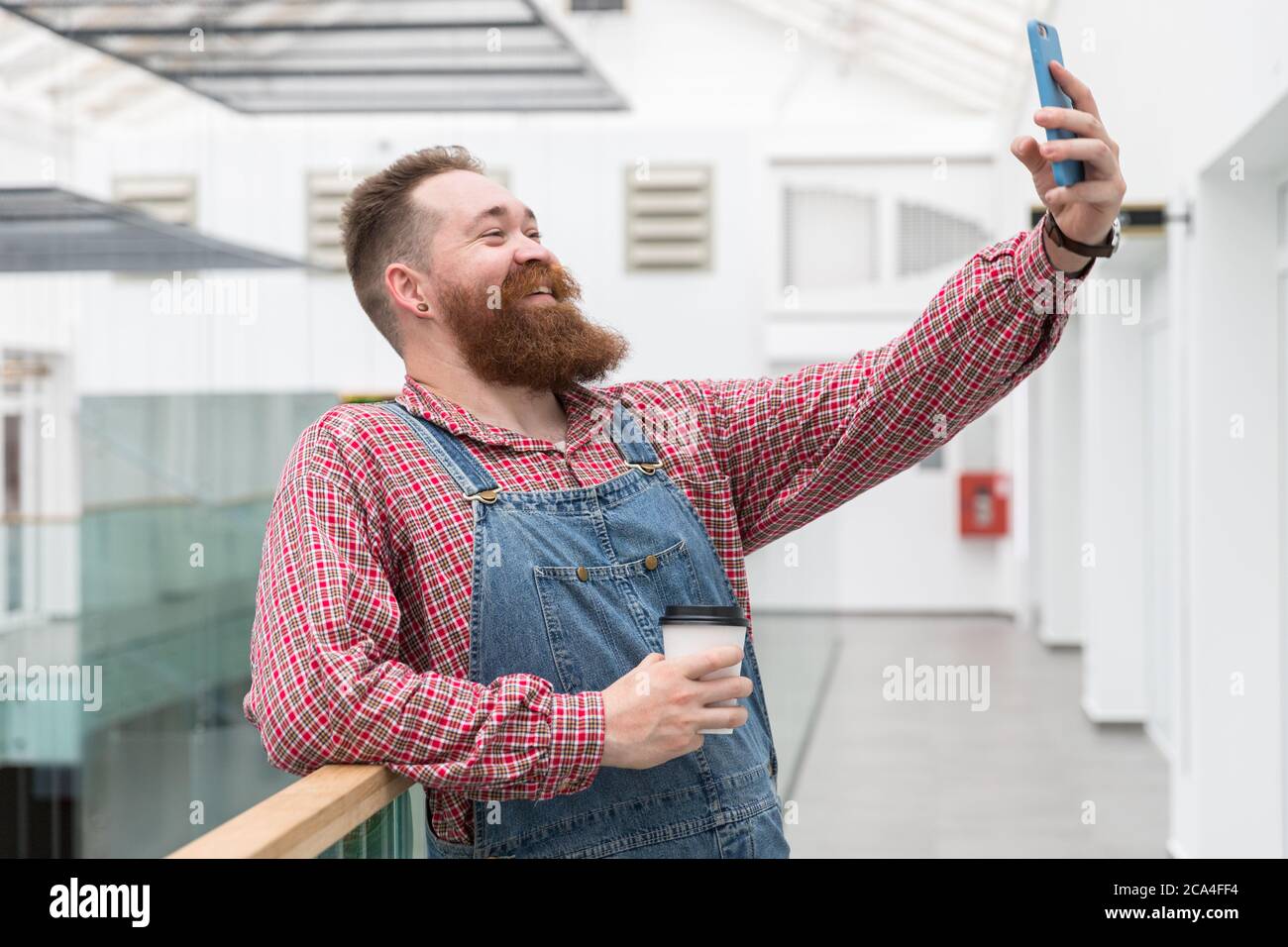 Sorridente barbiere in tute blu, camicia controllata, caffè da una tazza di carta, parlare in videochiamata o prendere un selfie su smartph Foto Stock