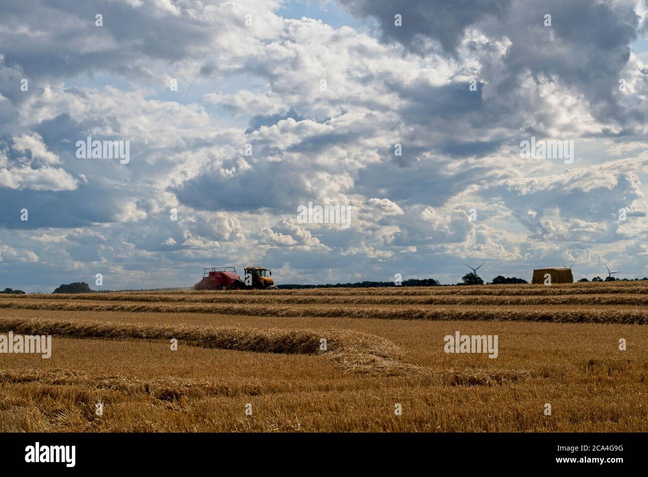 Imballatura dopo la mietitura vista a distanza del rimorchio del trattore che espelle la balla sul terreno cielo nuvoloso stoppie in primo piano che si muovono da sinistra a destra. Foto Stock