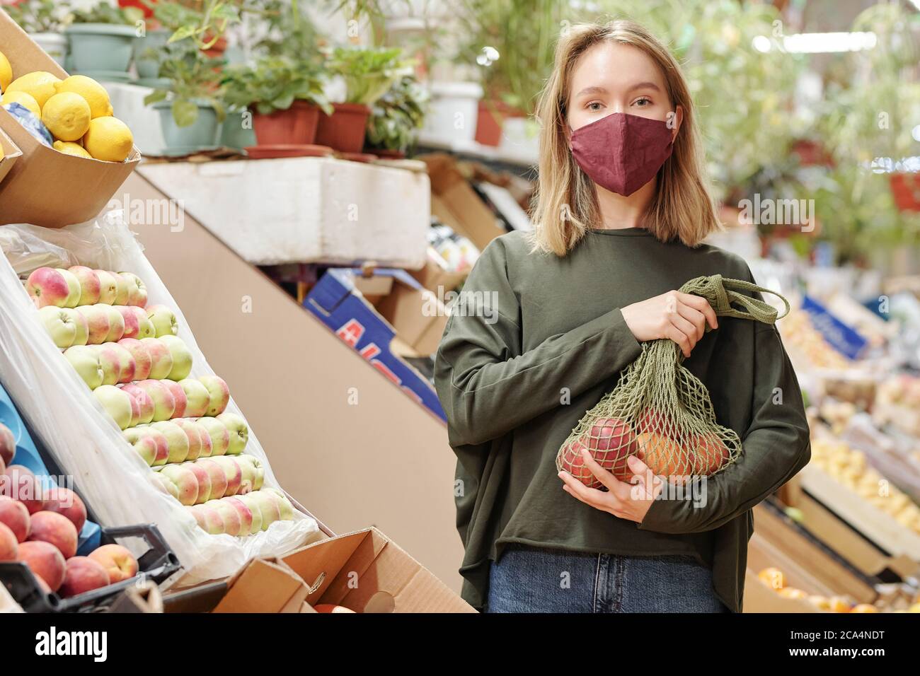 Ritratto di bella ragazza in maschera che tiene sacchetto di rete di prodotti biologici al mercato degli agricoltori durante il coronavirus Foto Stock