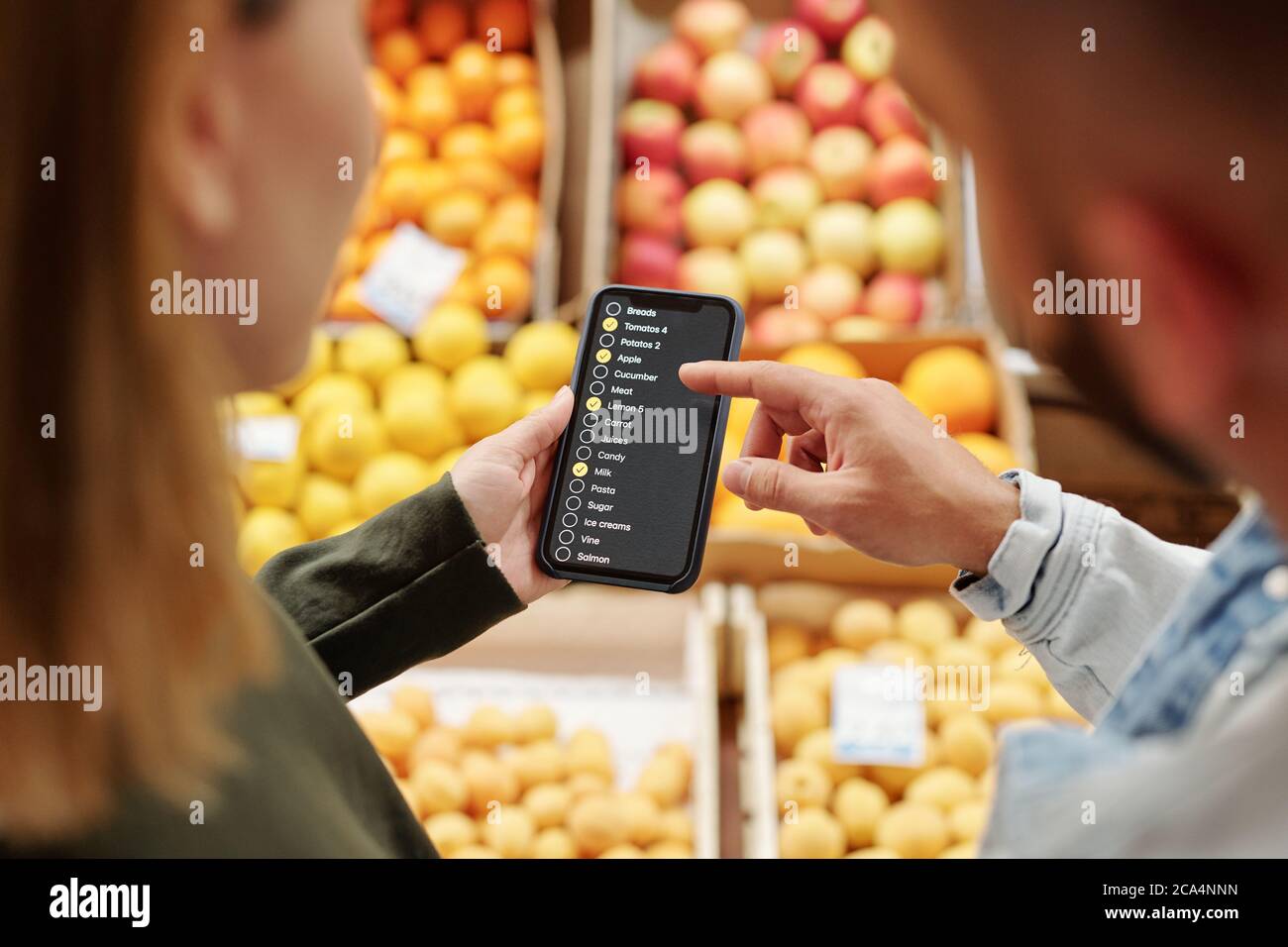Vista a spalla della giovane coppia che controlla l'elenco dei prodotti sullo smartphone durante l'acquisto di prodotti alimentari al mercato agricolo Foto Stock