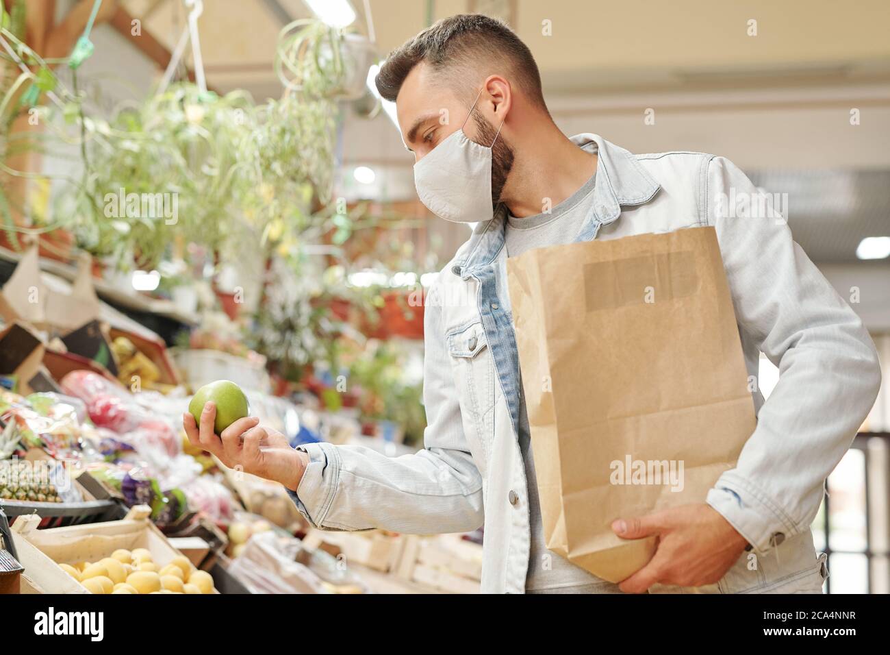 Giovane uomo in maschera facciale che tiene il sacchetto di carta e la scelta della frutta mentre comprando i generi alimentari freschi al mercato organico Foto Stock