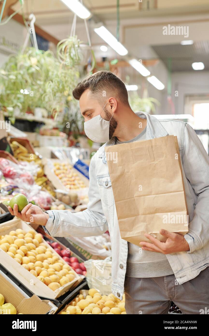 Giovane cliente maschio in maschera di stoffa in piedi al bancone del cibo e tenendo in mano il sacchetto di carta mentre comprando le mele al mercato degli agricoltori Foto Stock