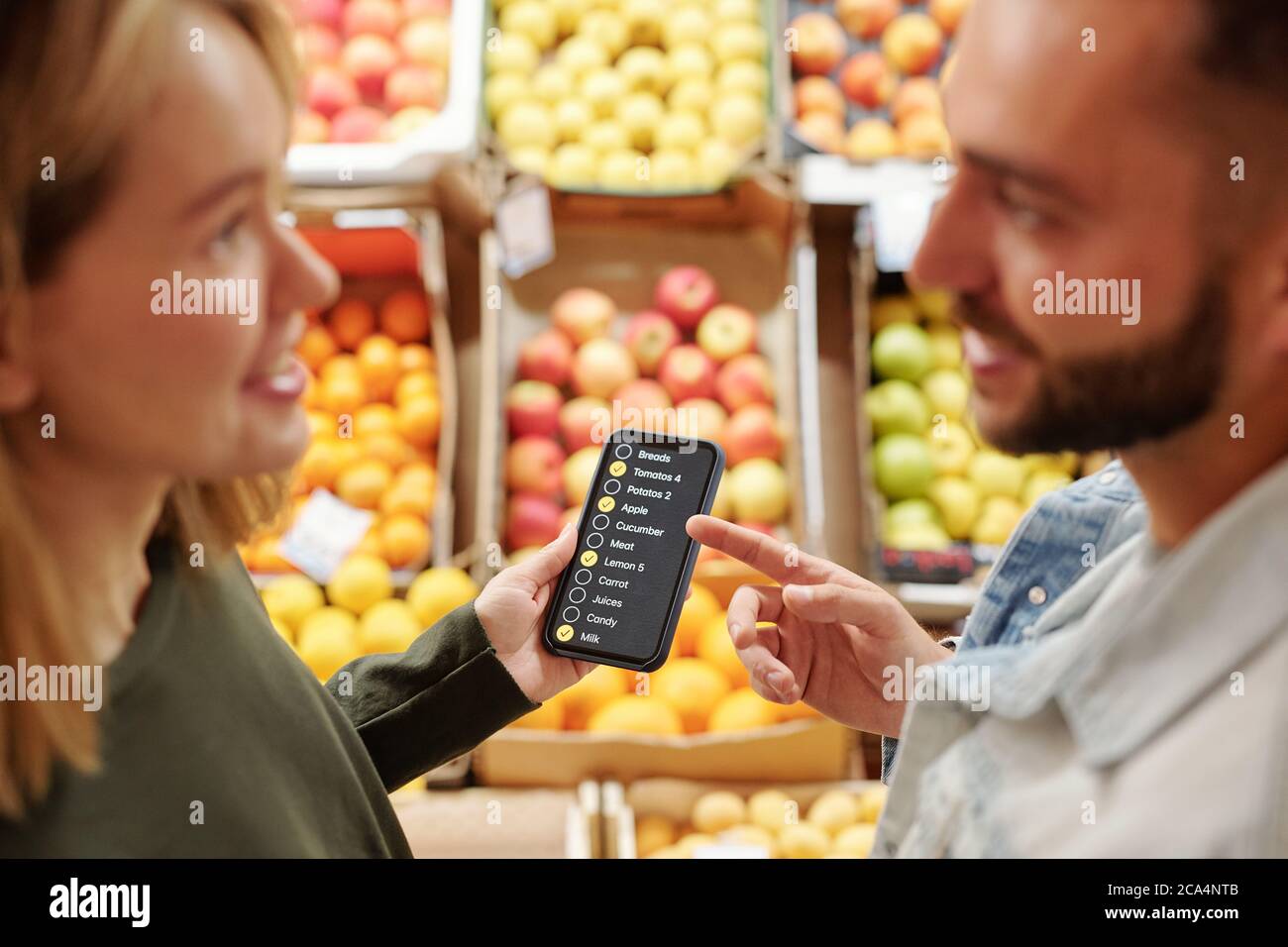 Una giovane coppia positiva che si trova al bancone del cibo e utilizza l'app per smartphone mentre discute l'elenco dei prodotti sul mercato biologico Foto Stock