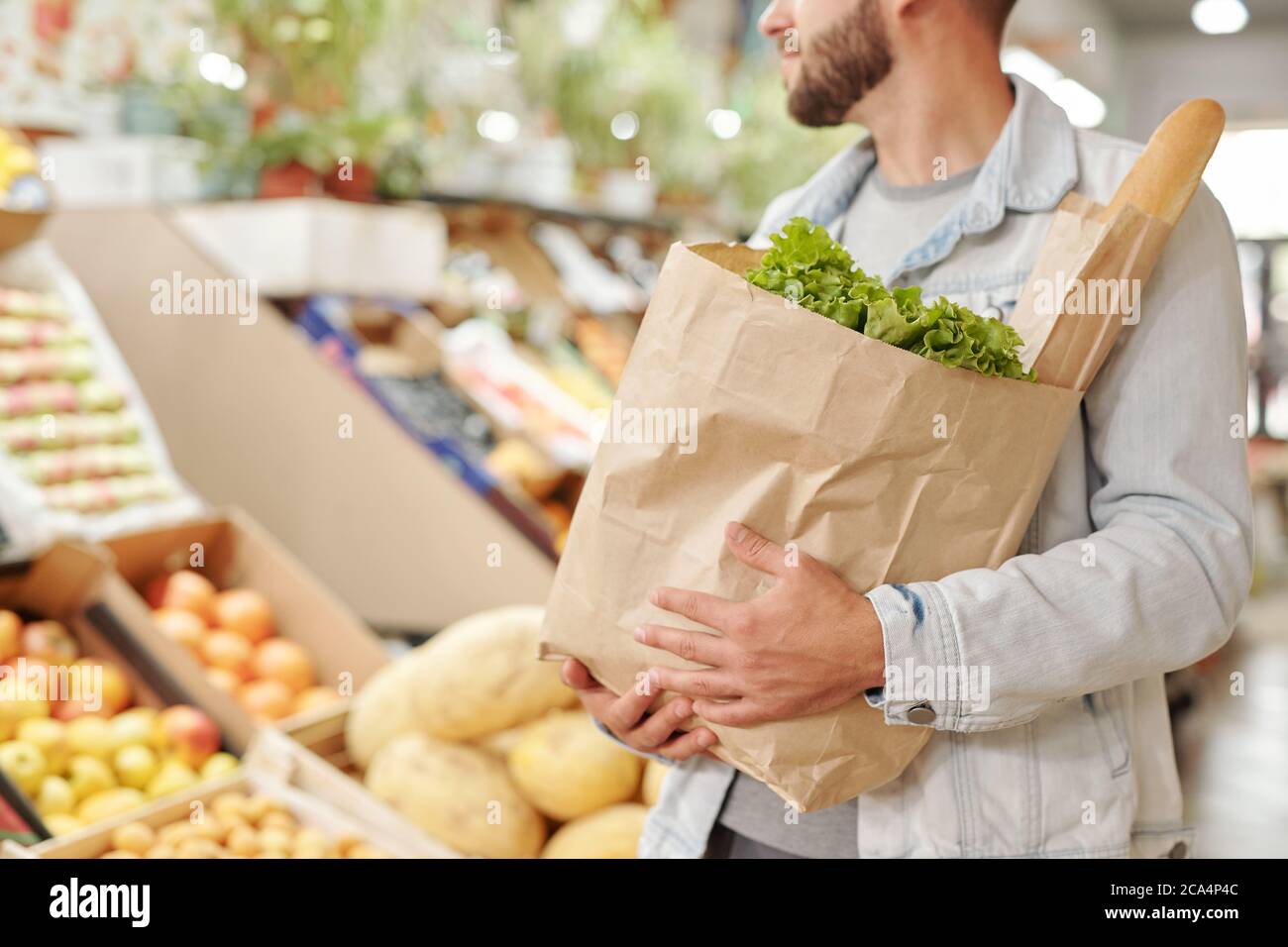 Primo piano di uomo bearded in giacca di denim che trasporta il sacchetto pieno di carta dei prodotti freschi al mercato alimentare Foto Stock