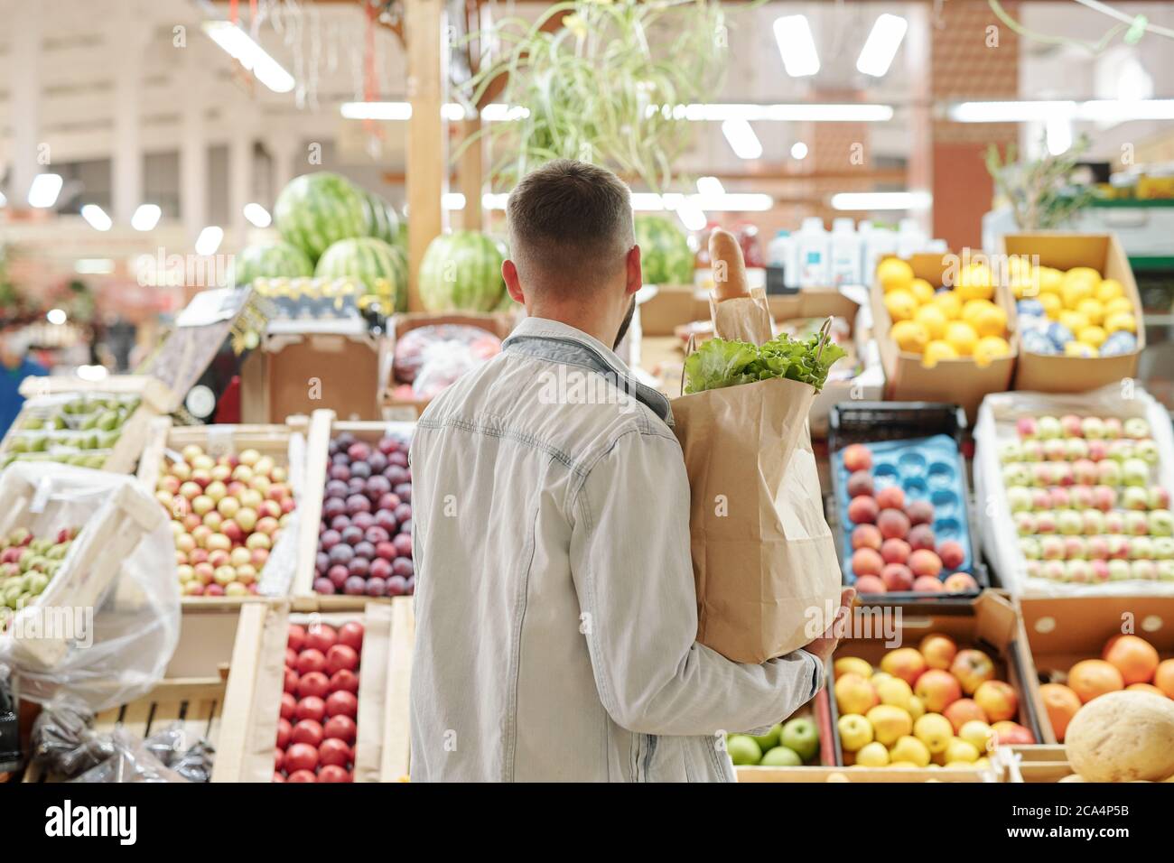Vista posteriore del giovane uomo con giacca in denim leggero che tiene il sacchetto di carta e la scelta della frutta succosa al mercato agricolo Foto Stock