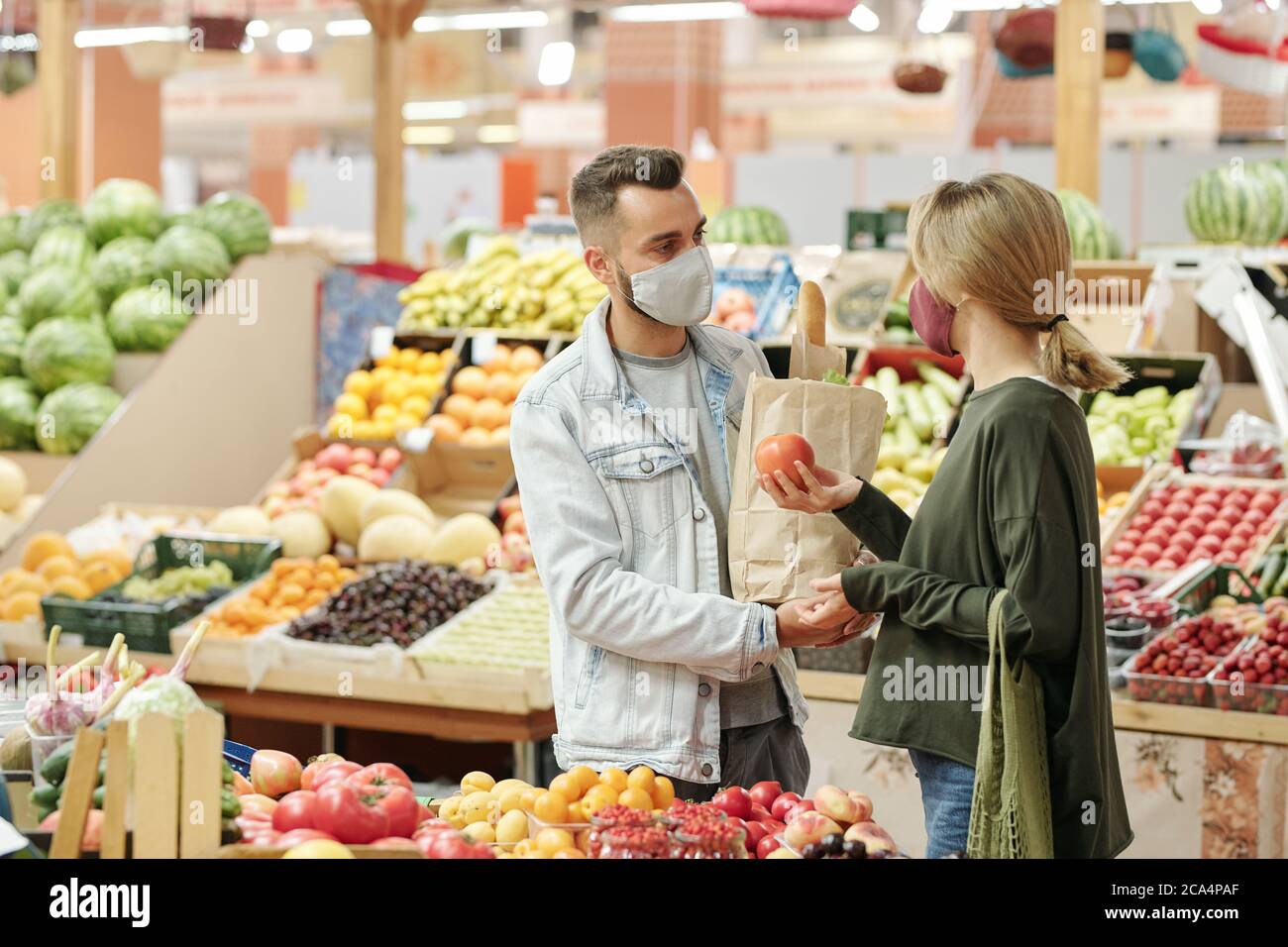Giovane coppia in maschere di stoffa in piedi al banco e l'acquisto di cibo organico al mercato: Ragazza che sceglie i pomodori Foto Stock