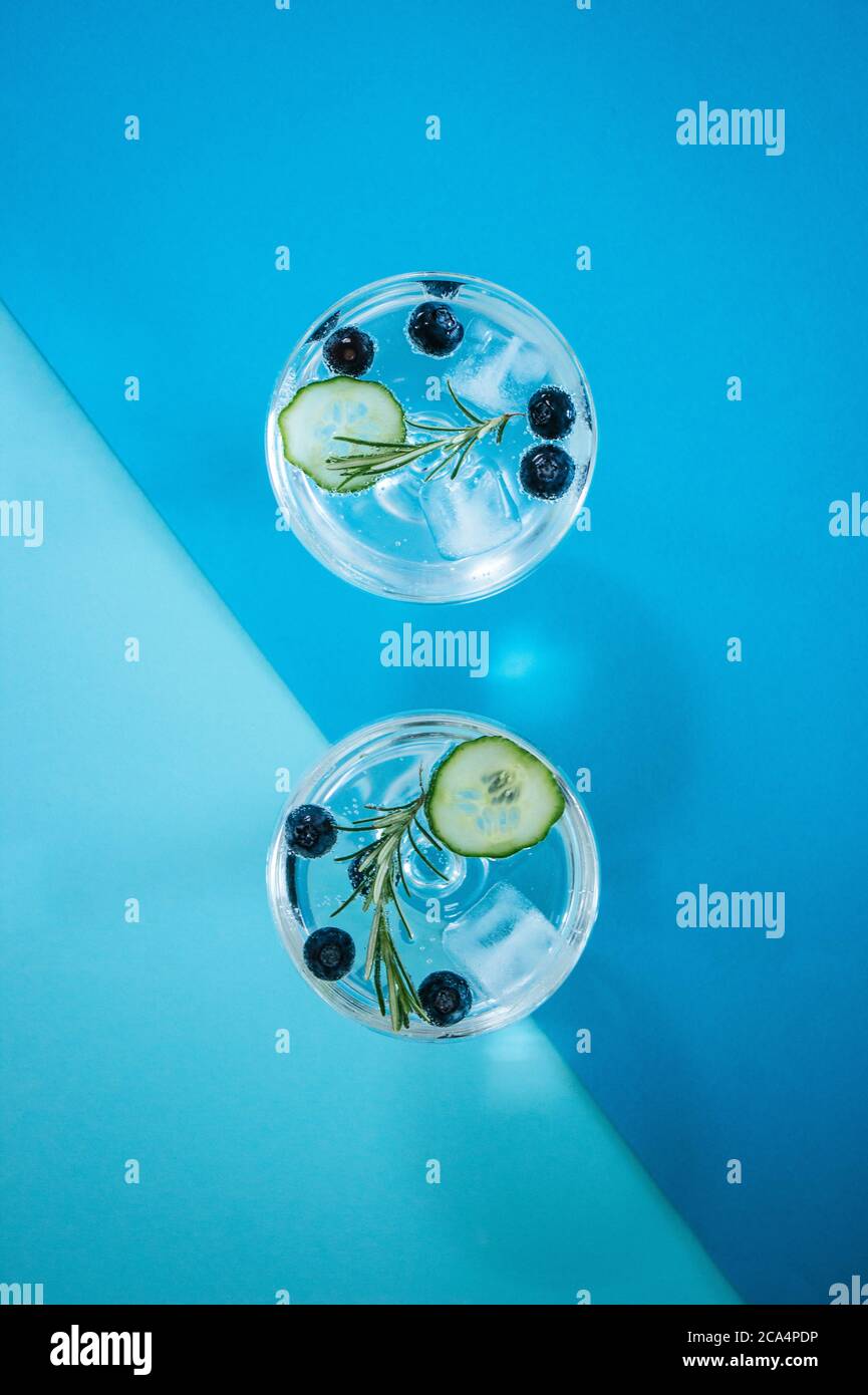 Due bicchieri di gin e cocktail tonico con mirtilli, cetrioli e rosmarino isolati su uno sfondo astratto e geometrico di colore blu Foto Stock