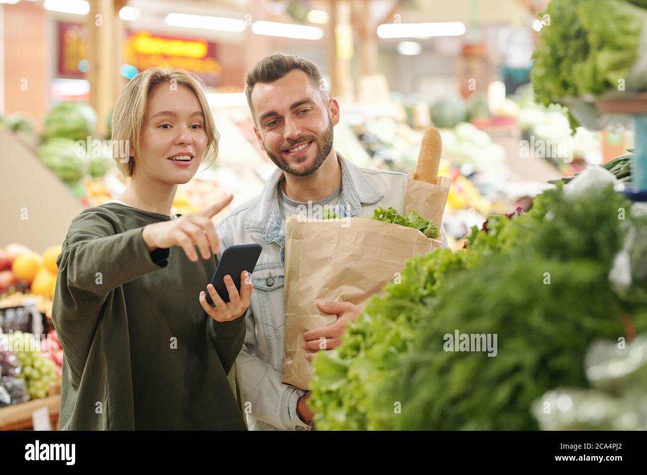 Una giovane donna positiva che utilizza l'app per smartphone per acquistare e indicare i green al ragazzo mentre acquista prodotti sul mercato Foto Stock