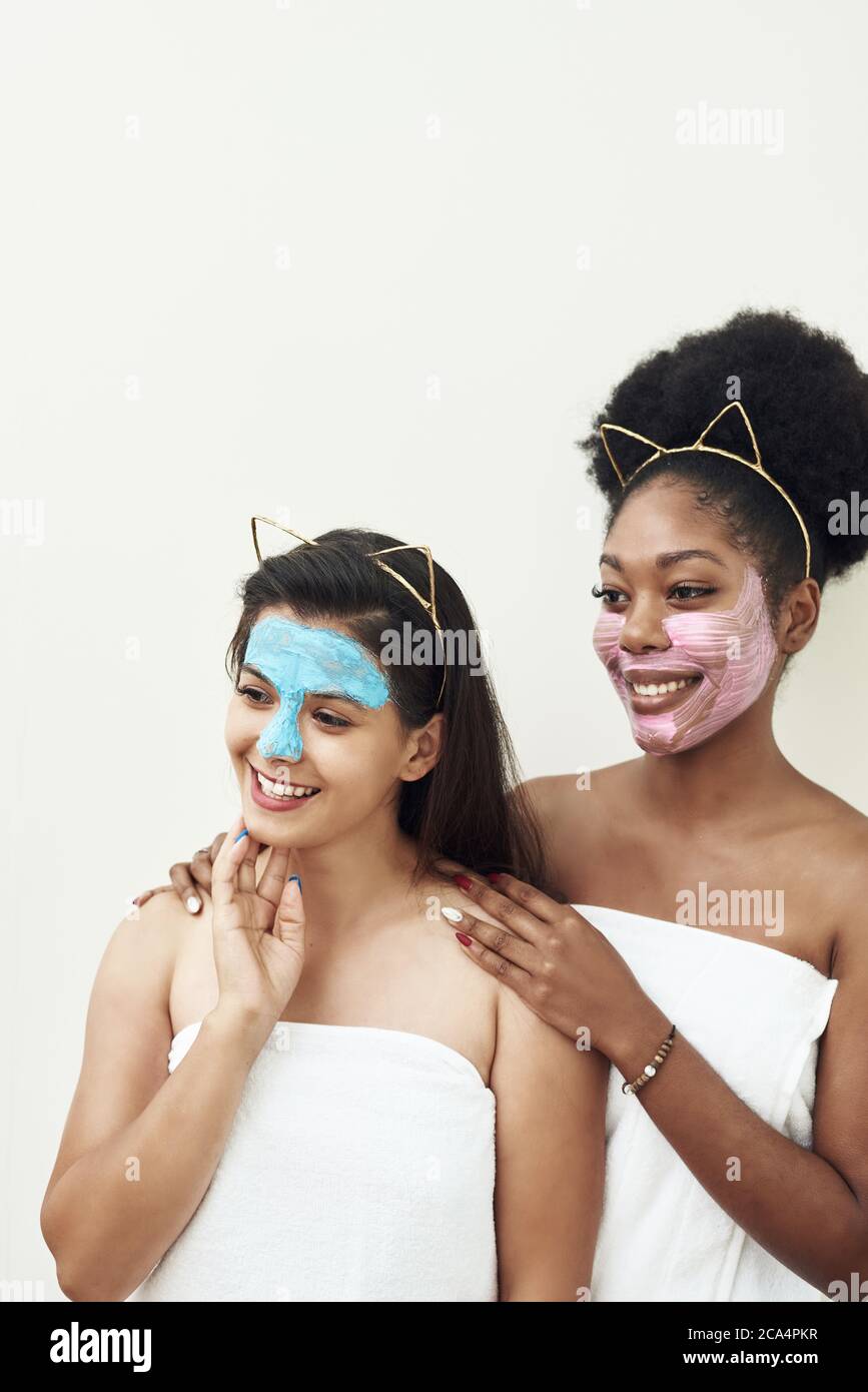 Multimasking. Due diverse ragazze etniche sono abbraccianti. I volti delle ragazze sono ricoperti di maschere per la cura del viso rosa e blu. Bellezza viso, trattamento viso e concetto spa. Foto Stock