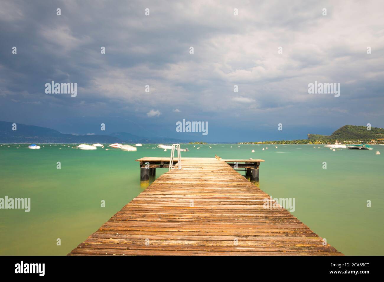 Un ponte pedonale conduce al tormentato Lago di Garda in Italia. La superficie blu-verde dell'acqua è orlata, un sacco di onde. Le barche ballano in acqua. Il cielo è b Foto Stock