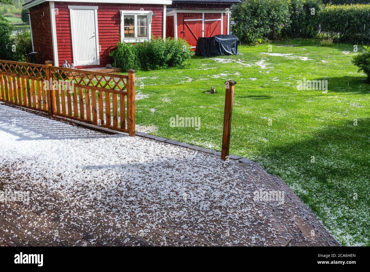Vista sulla terrazza in legno e sul giardino verde con pietre di grandine durante la tempesta dal cielo con luce solare Foto Stock