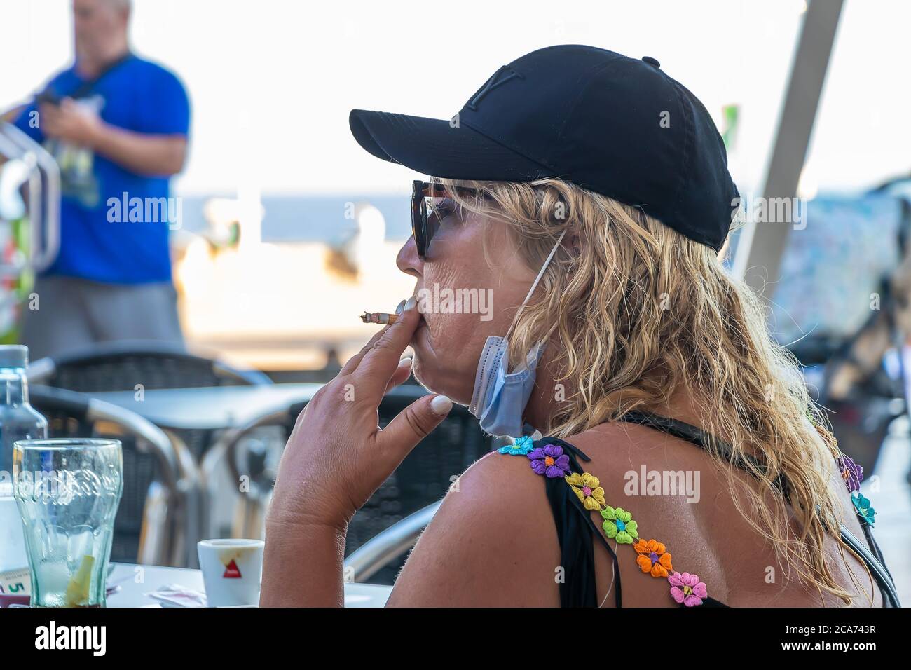 Huelva, Spagna - 10 luglio 2020: Donna bionda matura che indossa la maschera facciale per evitare il coronavirus covid-19 è fumare una sigaretta in una terrazza bar Foto Stock