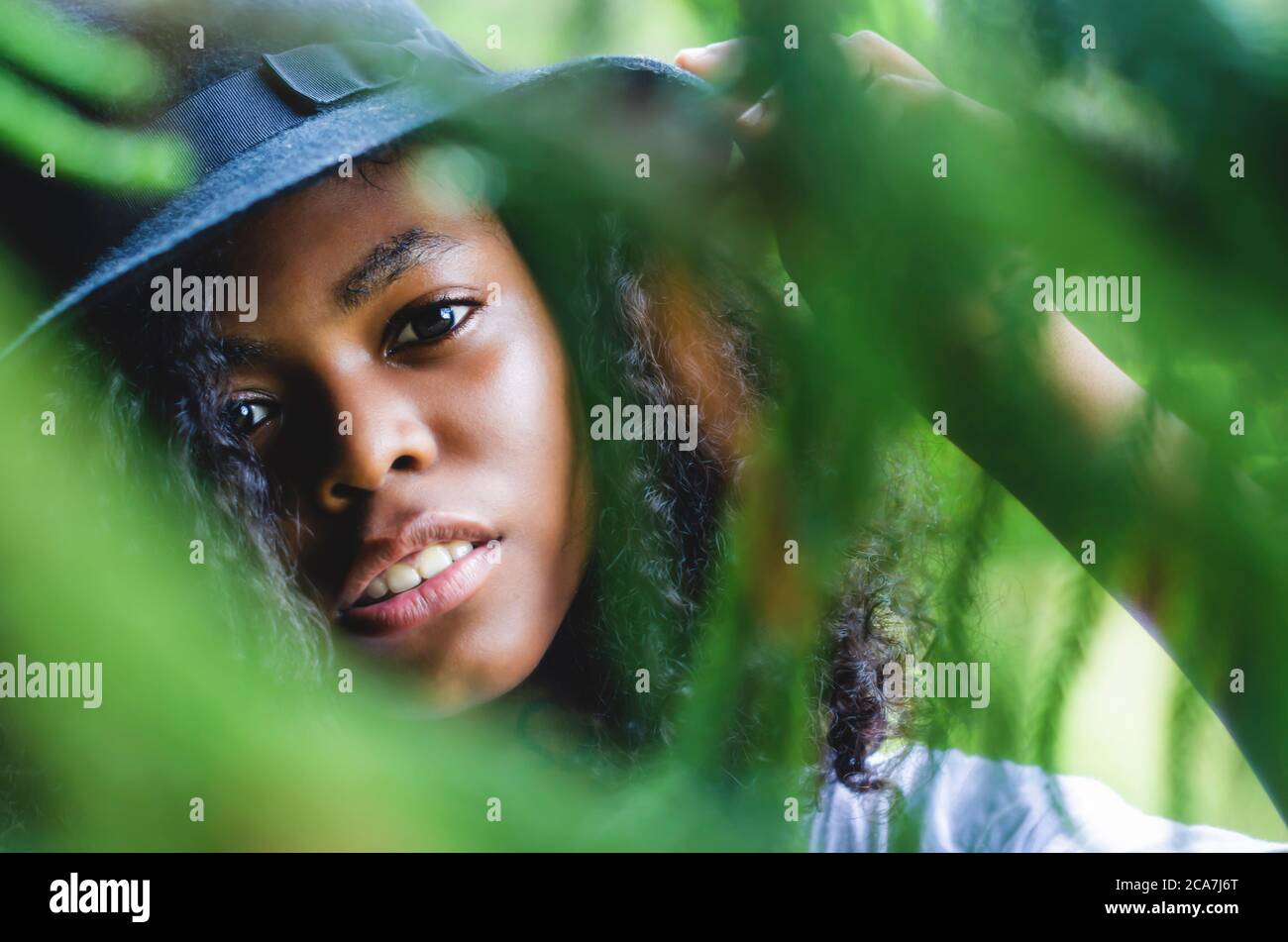 donna nera da 20 a 25 anni guardando la macchina fotografica attraverso la vegetazione, con cappello nero e capelli ricci Foto Stock