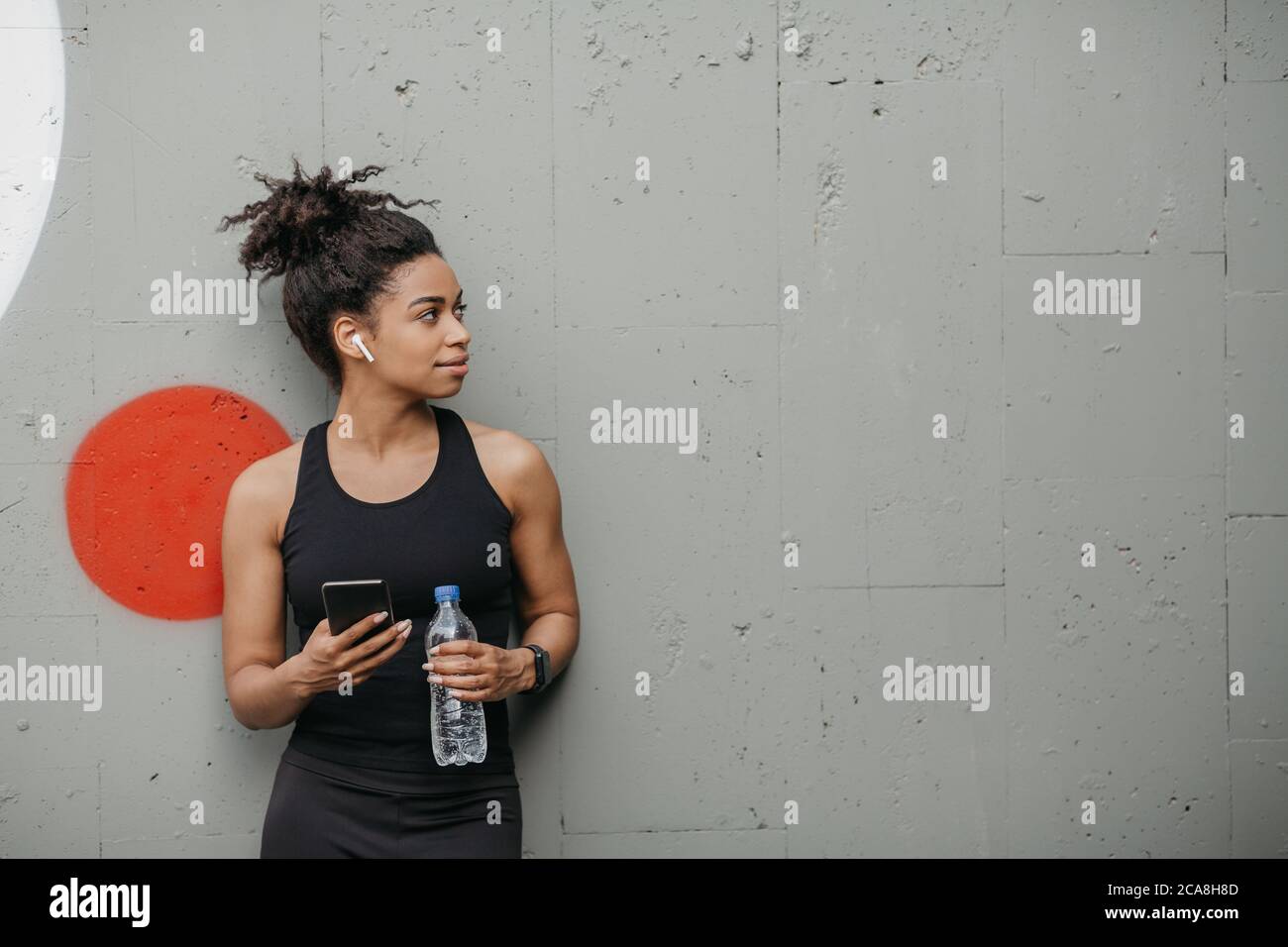 Muscolosa afroamericana ragazza con moderni gadget per l'allenamento tiene bottiglia e smartphone su sfondo grigio parete in città Foto Stock