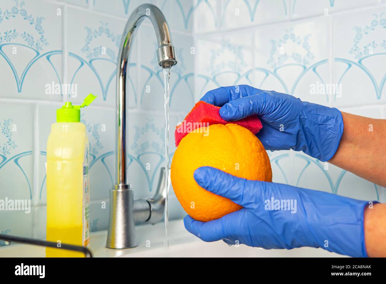 La persona lava la frutta con sapone ecologico per il lavaggio in una cucina con pareti blu. Decontaminazione e disinfezione dei prodotti dal punto vendita. Prevenzione di Foto Stock