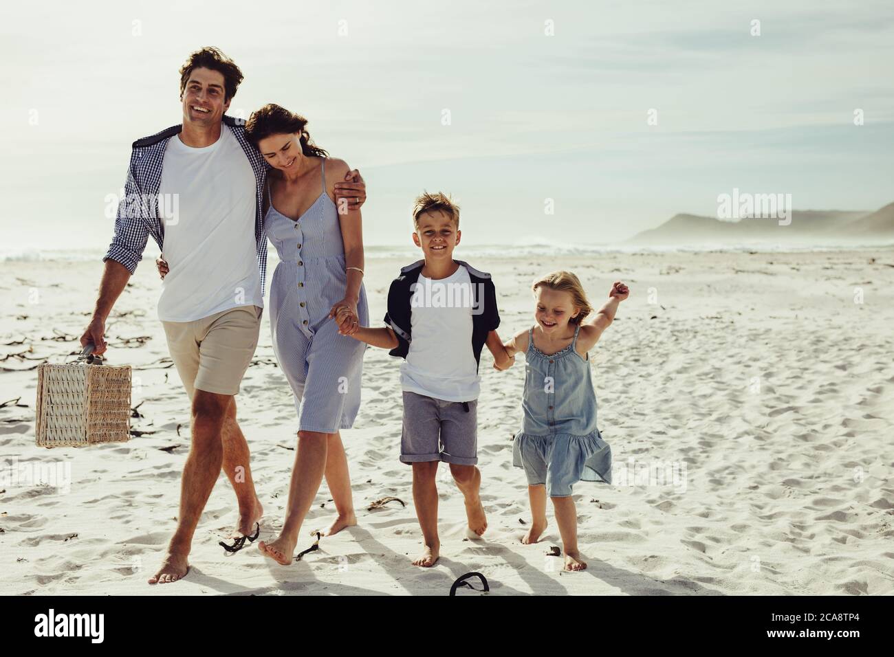 Bella famiglia di quattro in vacanza estiva in spiaggia. Famiglia che cammina insieme sulla spiaggia, con l'uomo che porta un cestino da picnic. Foto Stock