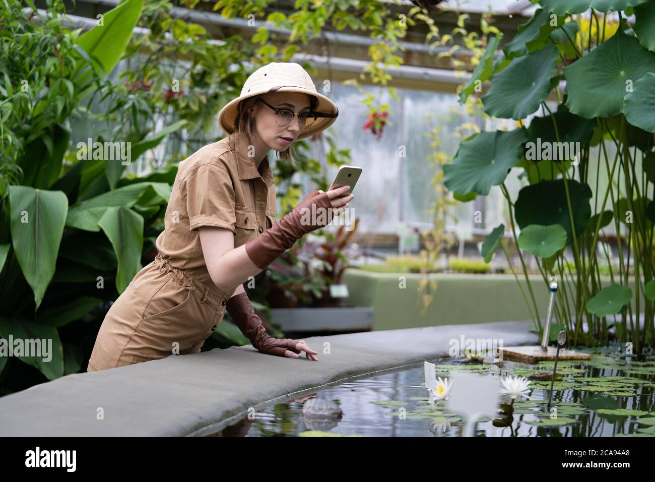 Donna botanica vestita in stile safari in serra. Naturalista in abiti cachi esplorando piante tropicali acquatiche in serra, scatta una foto su sm Foto Stock