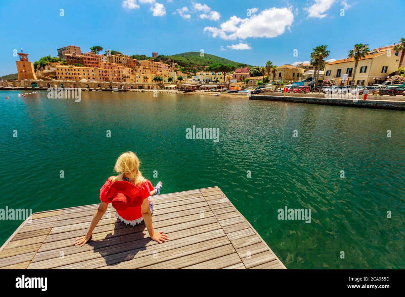 Donna bionda in tuta rossa che riposa e prende il sole sul molo nel porto di Rio Marina dell'isola d'Elba, Toscana, Italia, Europa Foto Stock