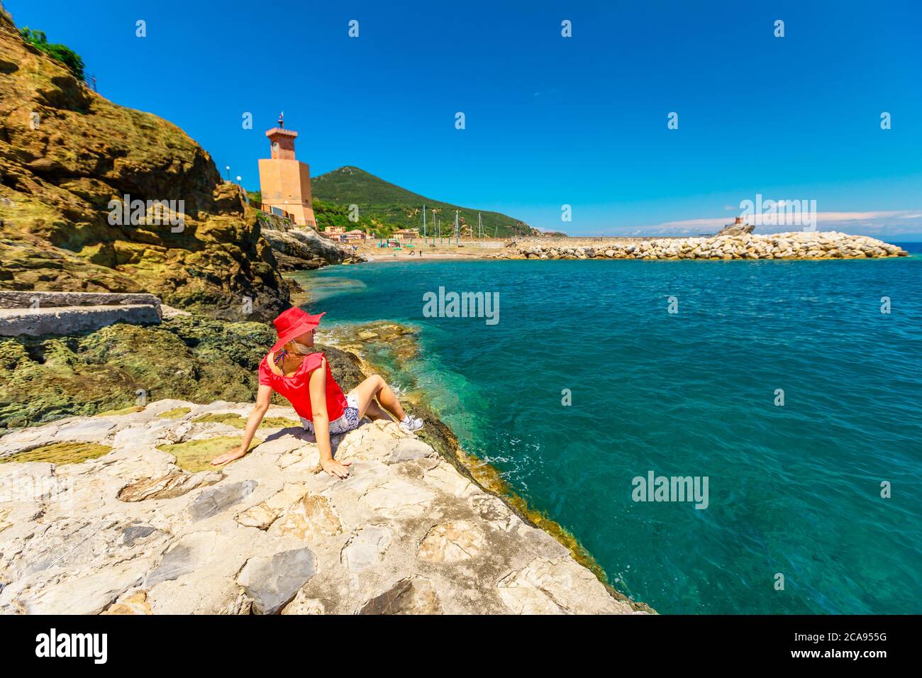 Felice donna in cappello rosso riposante sulle scogliere della spiaggia di Rio Marina, con torre dell'orologio sullo sfondo, Rio Marina, Isola d'Elba, Toscana, Italia, Europa Foto Stock