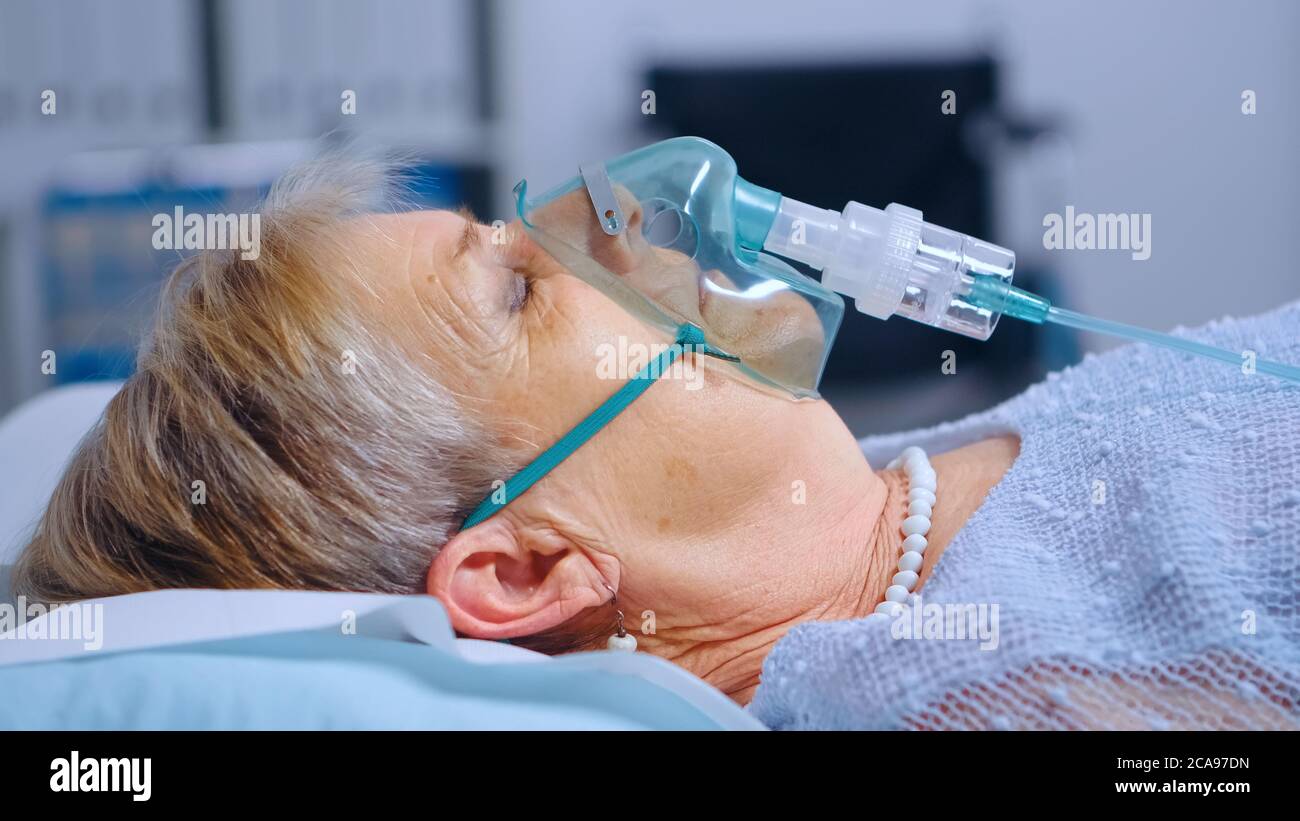 Primo piano ritratto di donna in pensione che respira con difficoltà nella maschera di ossigeno che si trova nel letto dell'ospedale durante l'epidemia di coronavirus covid-19. Medicina medica sistema sanitario. Trattamento delle infezioni Foto Stock