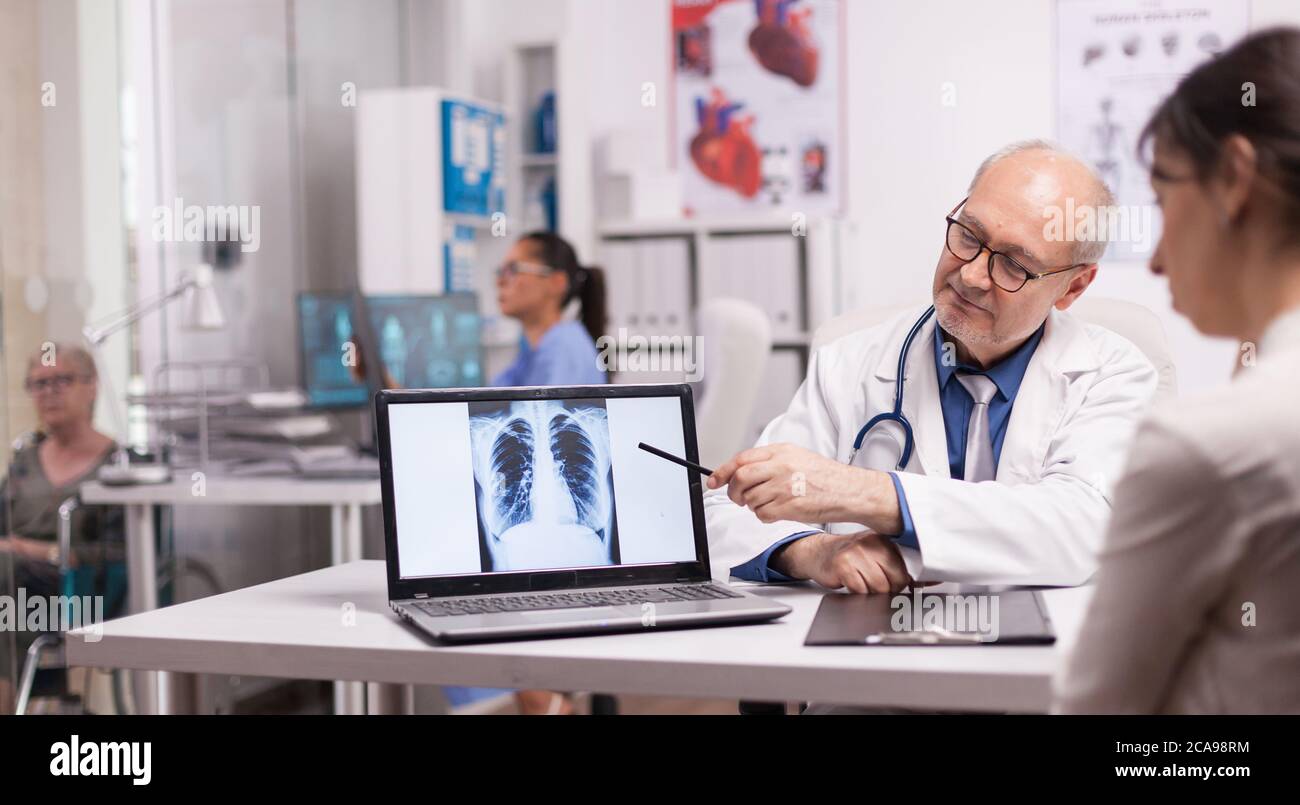 Medico senior che punta alla radiografia polmonare su un computer portatile per i pazienti giovani in ospedale. Donna disabile matura in sedia a rotelle che discute con medic circa la malattia. Foto Stock
