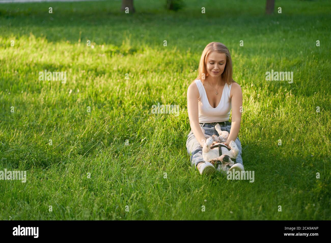 Vista frontale della giovane donna seduta con il bulldog francese maschile sulle gambe sull'erba. Attraente felice caucasica sorridente proprietario di cane femmina godendo il tramonto estivo con animale domestico, accarezzando il suo ventre nel parco della città. Foto Stock