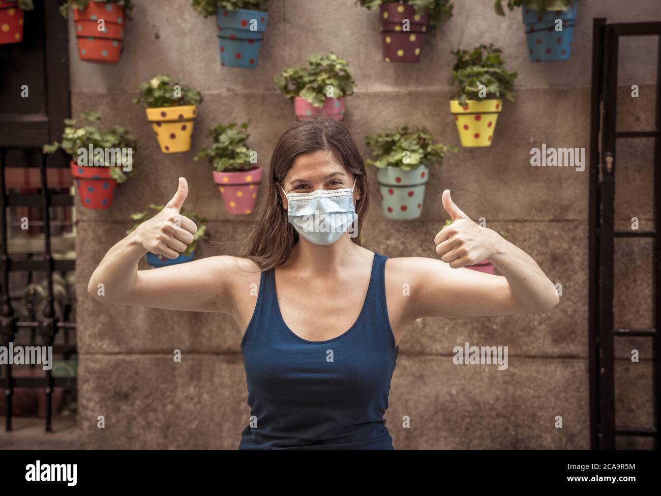 Giovane donna che indossa una maschera chirurgica sul viso mostrando i pollice su felice di essere all'aperto. Maschera di protezione da virus influenzali per la diffusione del coronavirus. Pos Foto Stock