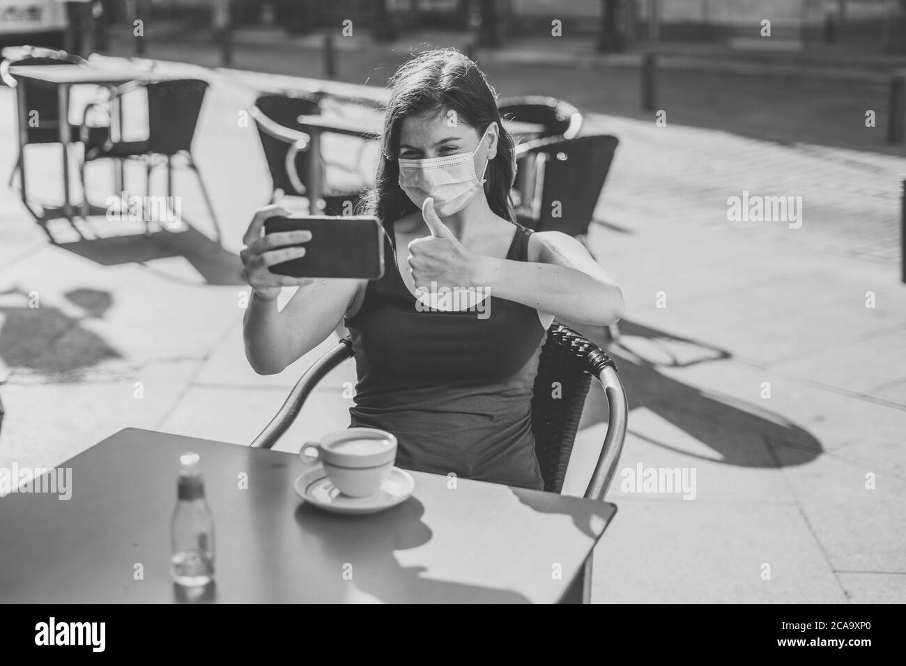 COVID-19 focolaio. Giovane donna che indossa una maschera chirurgica protettiva per il viso e che utilizza il telefono cellulare mostrando i pollici in su gesto in una terrazza bar. Corona Foto Stock