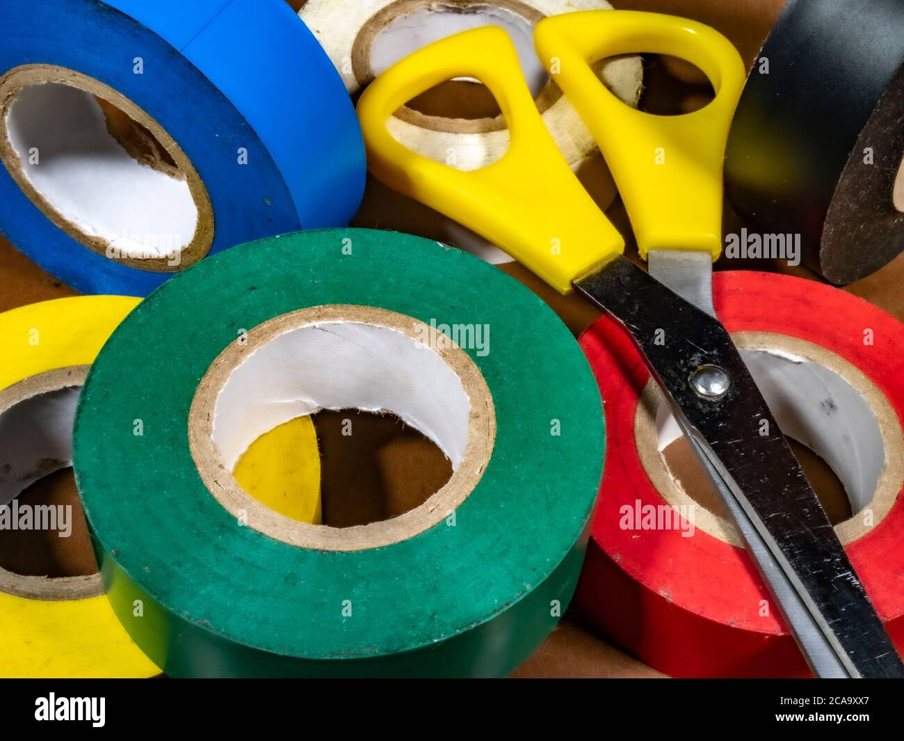 Primo piano POV di sei rotoli di nastro adesivo per uso domestico/elettrico, in una varietà di colori, più un paio di forbici. Foto Stock