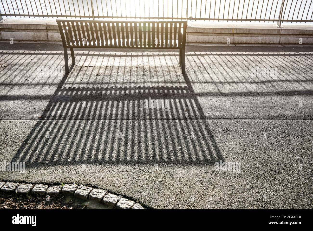 Mattina sole gettato linee di ombre su asfalto marciapiede attraverso recinzione e panca vuota vicino al fiume Foto Stock