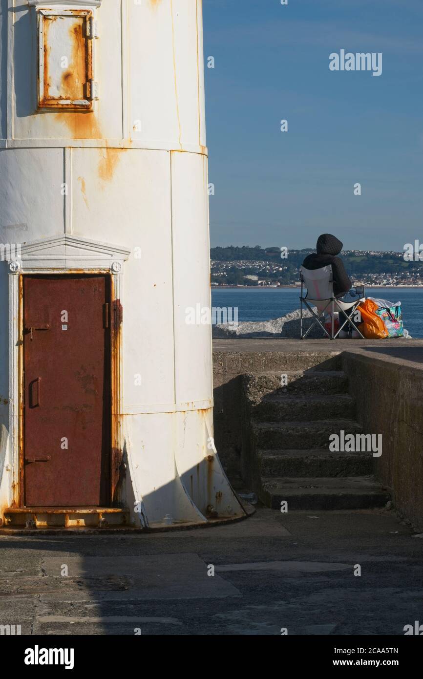 Il porto di Brixham si affaccia sul faro arrugginito alla fine del molo del porto. Vista della base e dei gradini che portano al livello superiore. Uomo seduto sulla sedia che guarda fuori. Foto Stock