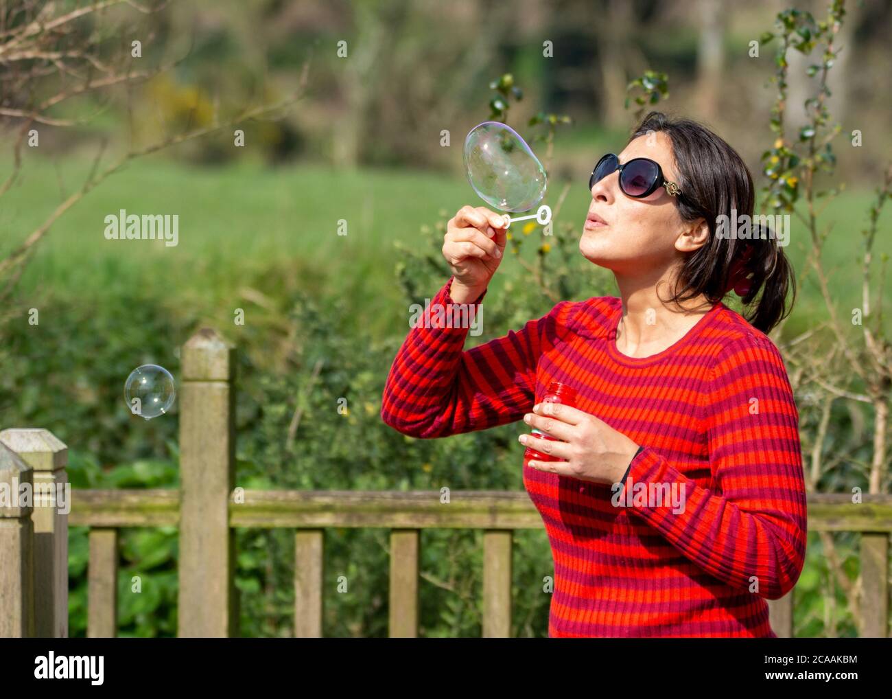 Donna con ponticello rosso, occhiali da sole e capelli neri lunghi che soffia bolle all'aperto Foto Stock