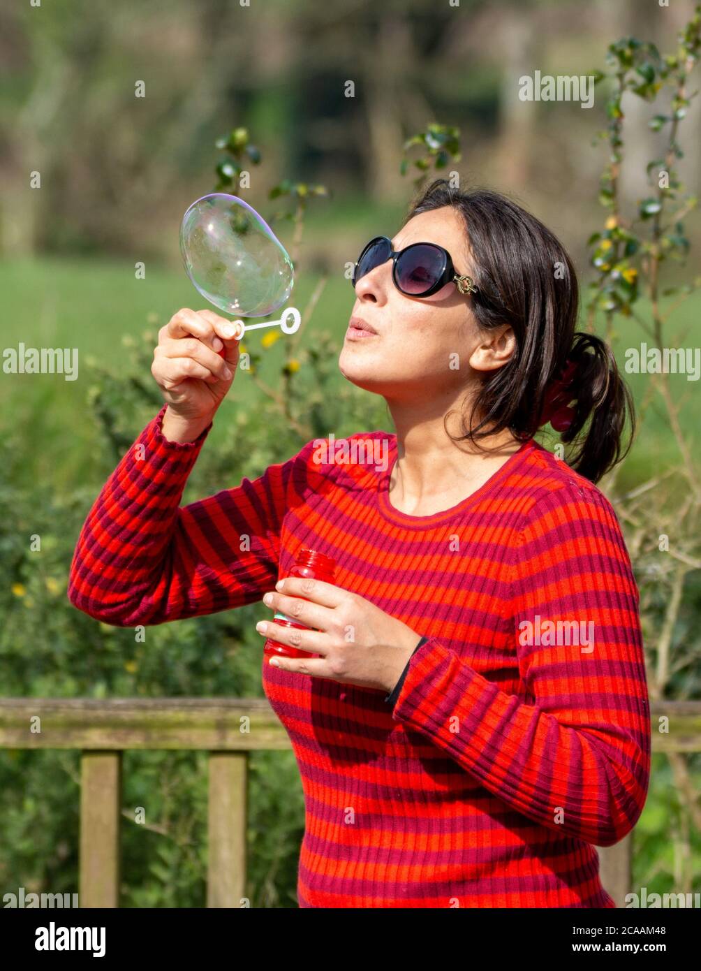 Donna con ponticello rosso, occhiali da sole e capelli neri lunghi che soffia bolle all'aperto Foto Stock
