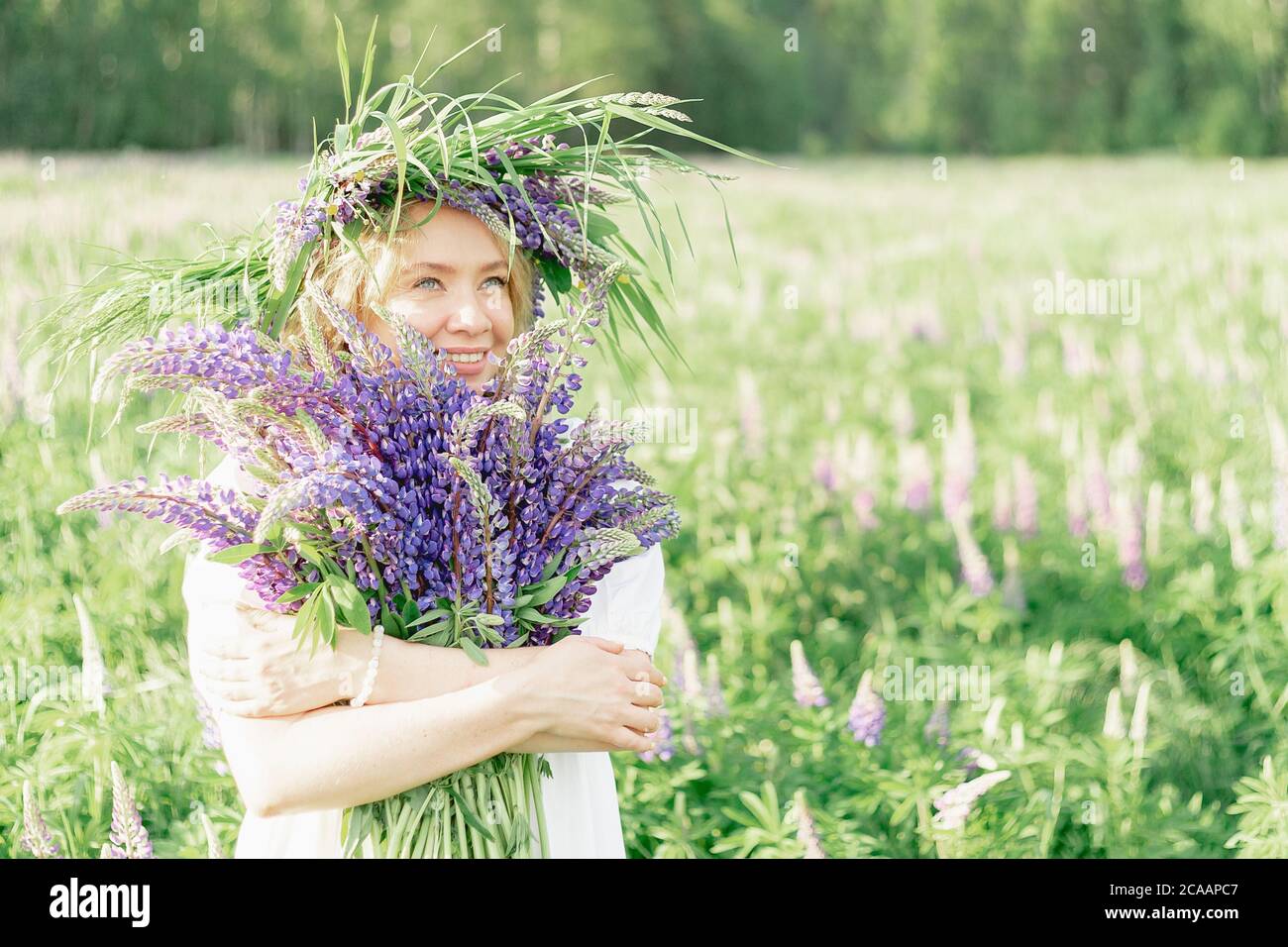 una ragazza felice in una corona di fiori abbraccia un bouquet di fiori selvatici e sorrisi, inala la fragranza di fiori, un bouquet di lupini. allegro allegro Foto Stock