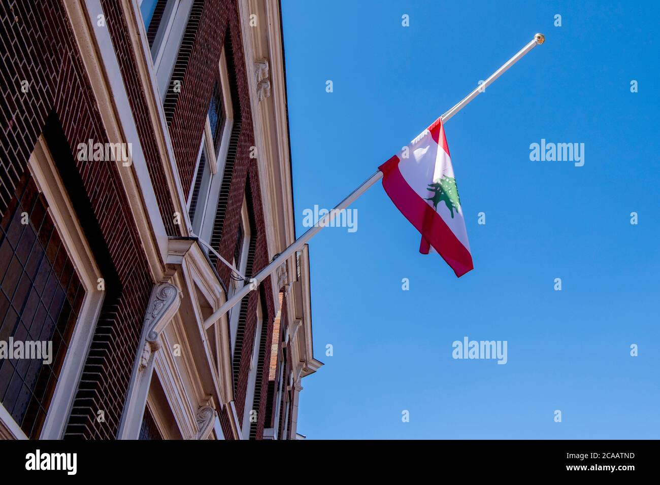 La bandiera del Libano è appesa a metà palo all'ambasciata del Libano dell'Aia dopo una potente esplosione che ha attraversato la capitale del Libano; Il risultato dell'accensione di un enorme deposito di nitrato di ammonio nel porto principale della città.i soccorritori hanno cercato i sopravvissuti a Beirut dopo un'esplosione cataclismica nel porto ha seminato devastazioni in interi quartieri, uccidendo più di 100 persone, provocando migliaia di morti e gettando il Libano in una crisi più profonda. L'esplosione, che sembrava essere stata causata da un incendio che infiamma 2,750 tonnellate di nitrato di ammonio lasciato incustodito in un magazzino, è stata avvertita lontano quanto il CYP Foto Stock