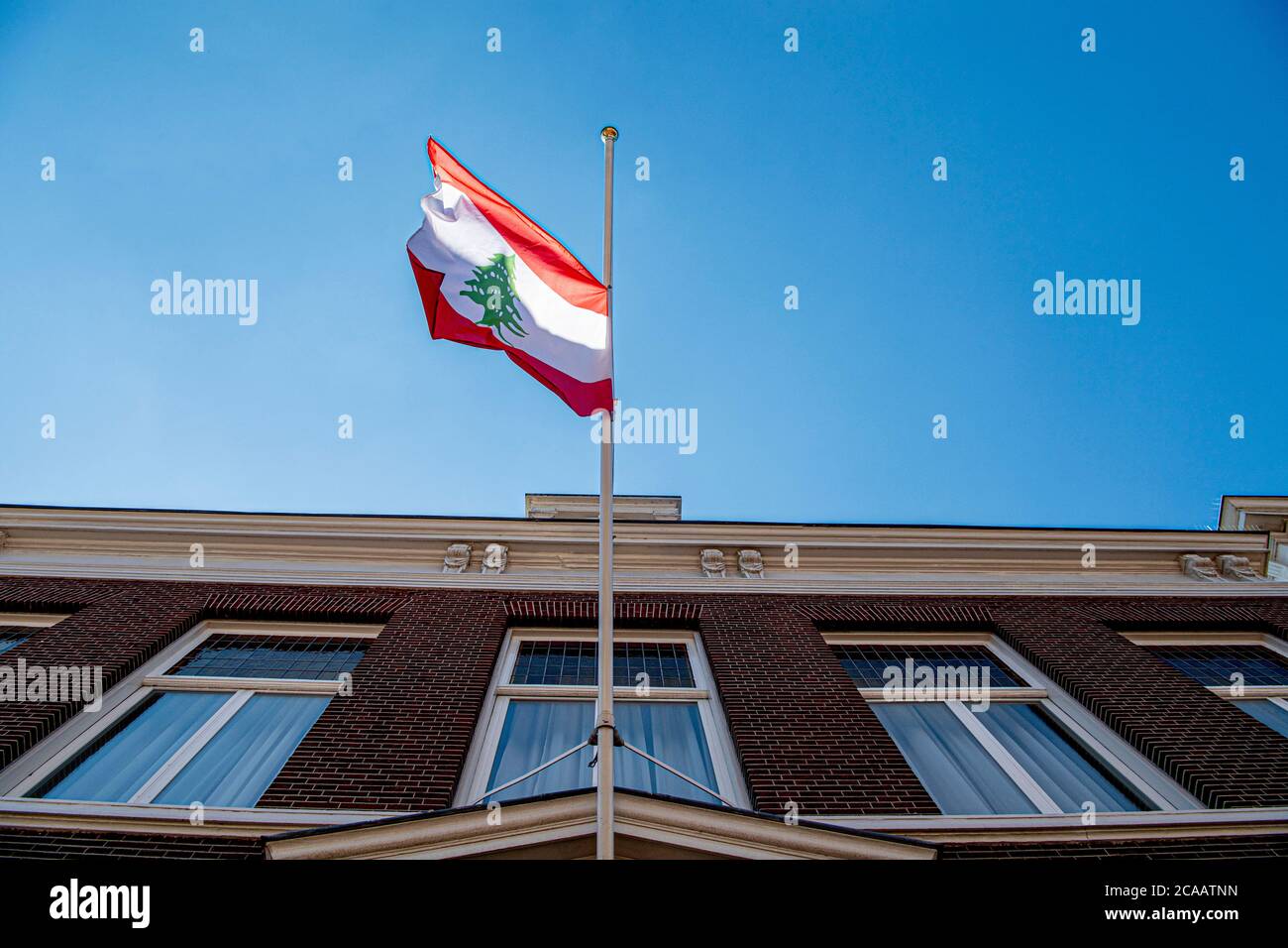 La bandiera del Libano è appesa a metà palo all'ambasciata del Libano dell'Aia dopo una potente esplosione che ha attraversato la capitale del Libano; Il risultato dell'accensione di un enorme deposito di nitrato di ammonio nel porto principale della città.i soccorritori hanno cercato i sopravvissuti a Beirut dopo un'esplosione cataclismica nel porto ha seminato devastazioni in interi quartieri, uccidendo più di 100 persone, provocando migliaia di morti e gettando il Libano in una crisi più profonda. L'esplosione, che sembrava essere stata causata da un incendio che infiamma 2,750 tonnellate di nitrato di ammonio lasciato incustodito in un magazzino, è stata avvertita lontano quanto il CYP Foto Stock