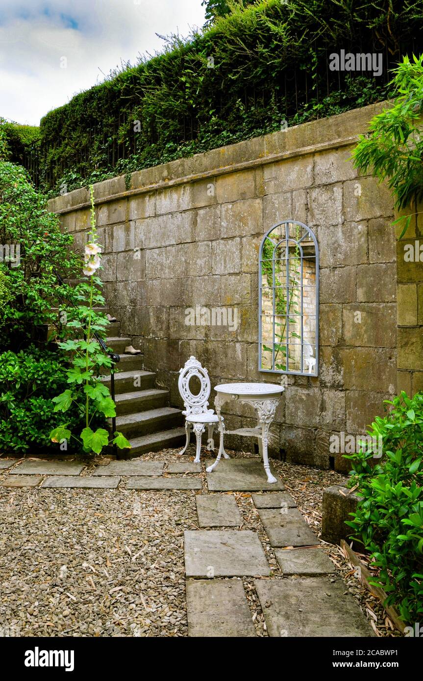 Un giardino cottage in pietra con mura, gradini, arbusti e arazzi. La parete è decorata con uno specchio georgiano davanti ad un ornato ferro battuto bianco Foto Stock