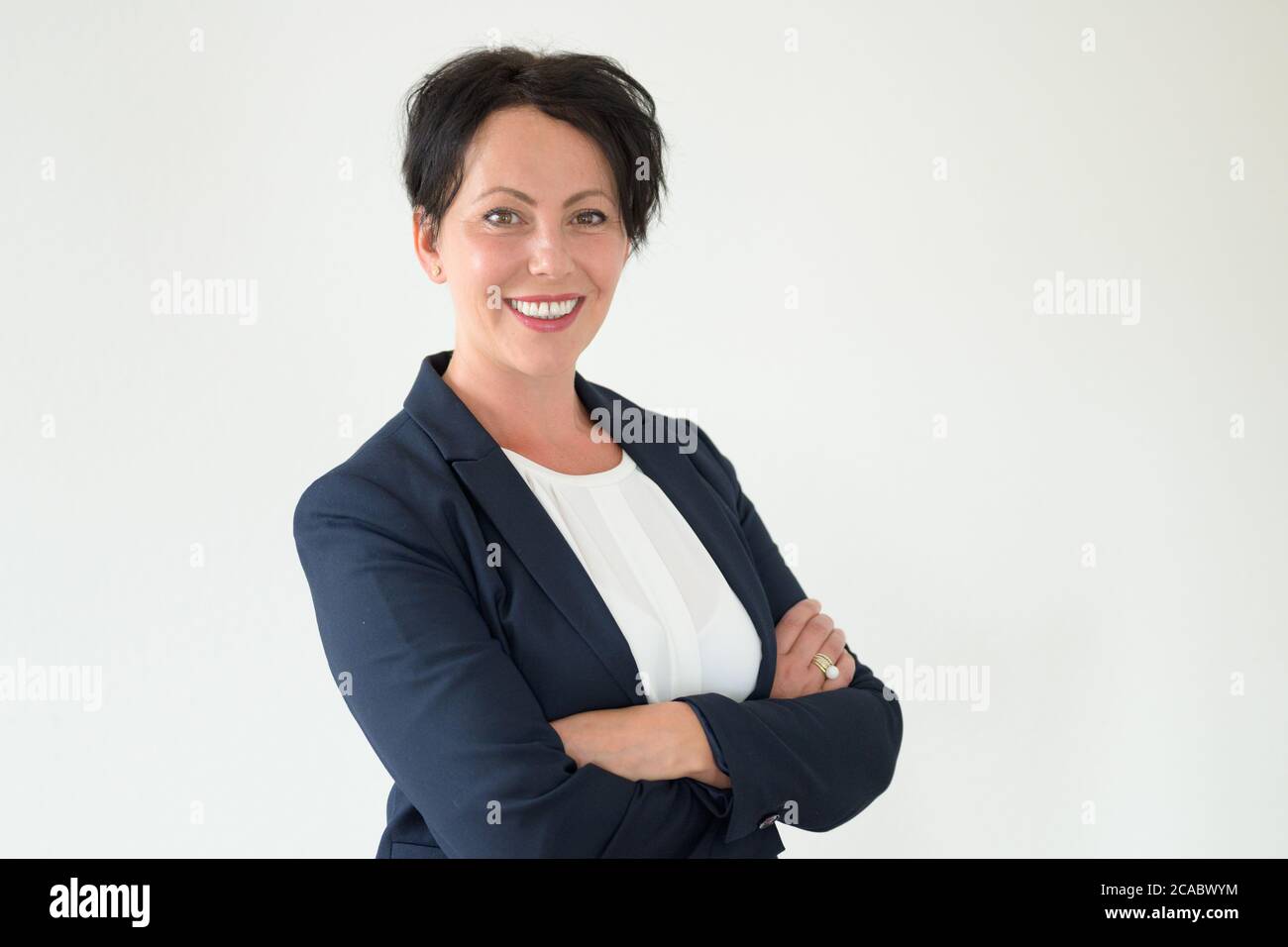 Fidata donna ispanica candidato di lavoro in giacca intelligente sorridente alla macchina fotografica come si alza con le braccia ripiegate contro una parete bianca con copyspace Foto Stock