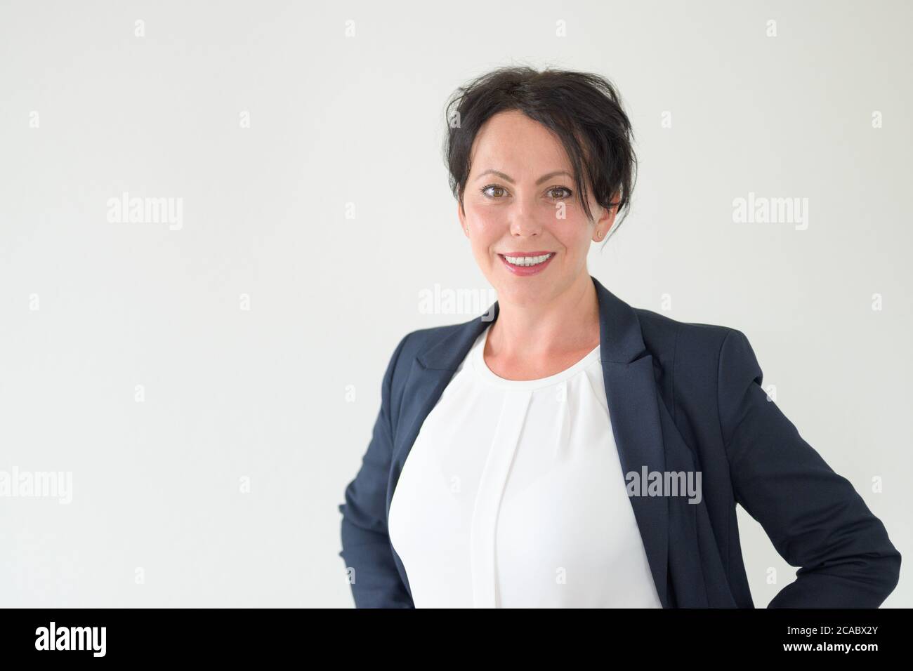 Donna d'affari ispanica attenta e sorridente, in piedi con le mani sui fianchi guardando la macchina fotografica in un concetto di candidato di lavoro contro una wa bianca Foto Stock