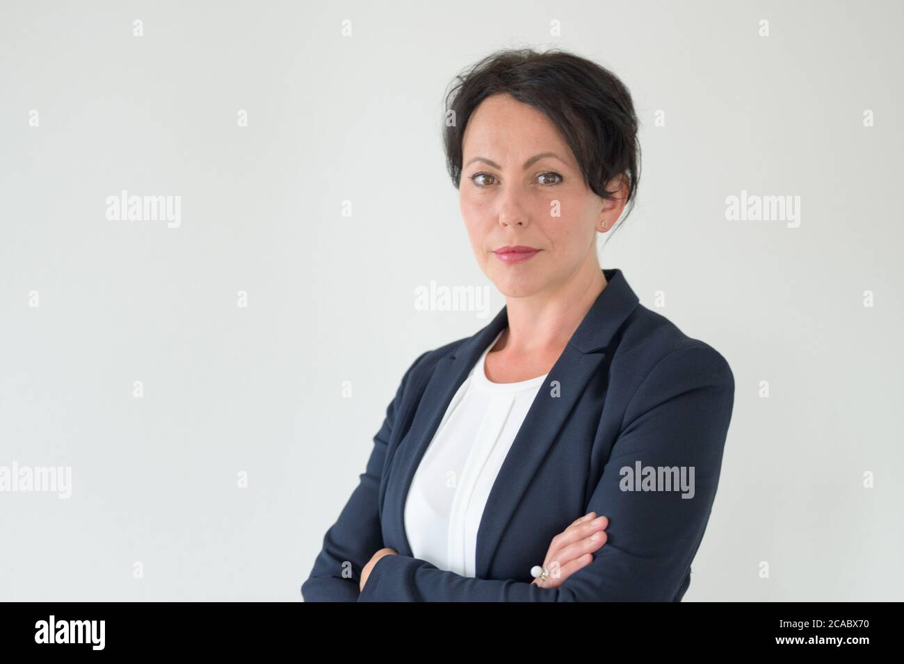 Donna d'affari pensiva seria fissando alla macchina fotografica con le braccia ripiegate in un ritratto di corpo superiore mezzo girato contro una parete bianca con copyspace Foto Stock