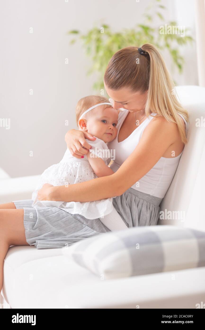 La mamma bionda giovane femmina tiene la sua figlia in braccio seduta su un divano a casa. Foto Stock